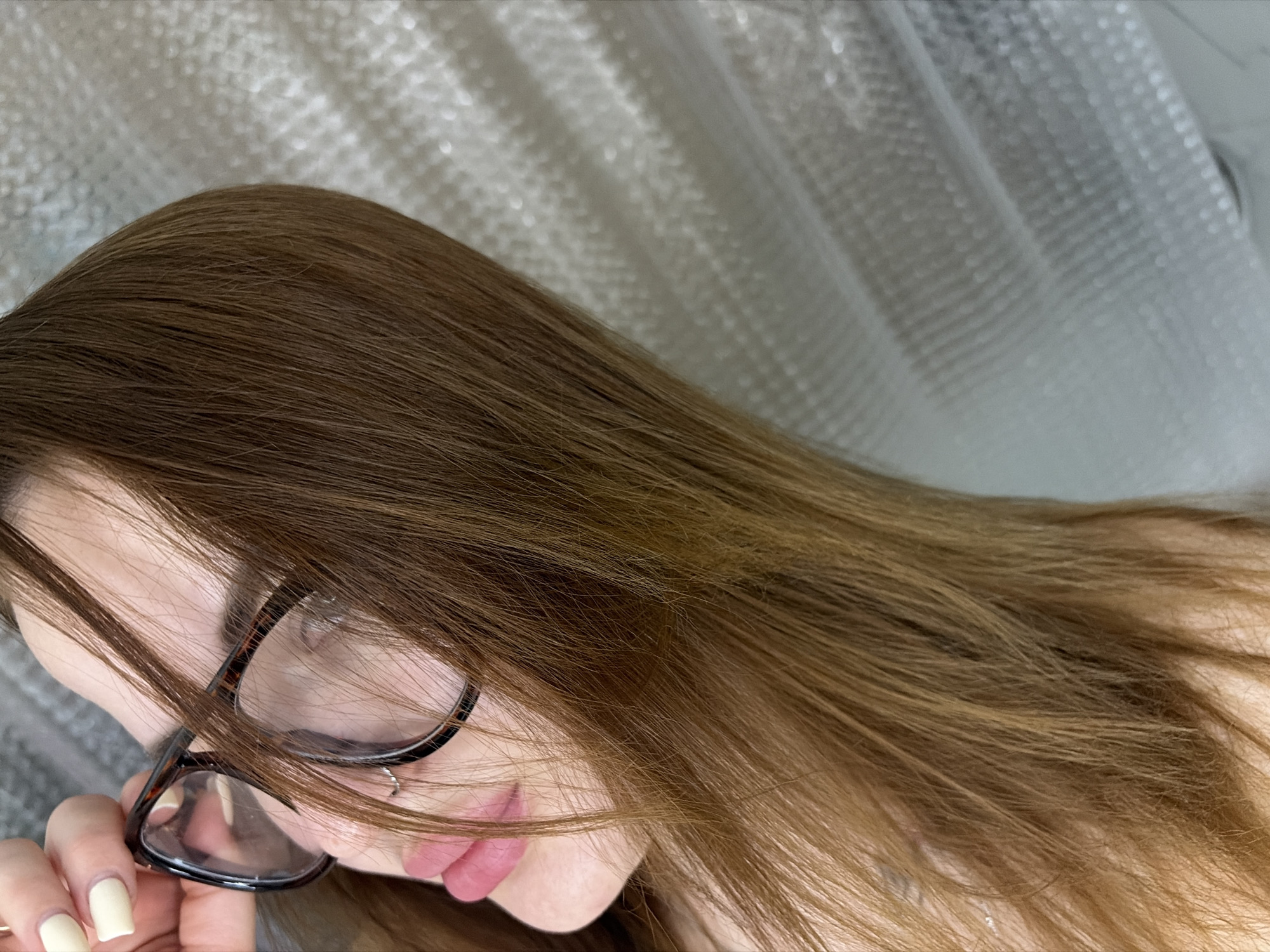 A close-up, angled portrait depicts a person with long, brown hair partially obscuring their face. The person wears glasses with dark frames. A small stud earring is visible in their nose. The person's hand, with fingernails painted in a pale yellow, holds the frame of the glasses. The background is a blurred, white, textured surface. The location is not discernible and cannot be determined to be in Krasnodar, Russia.