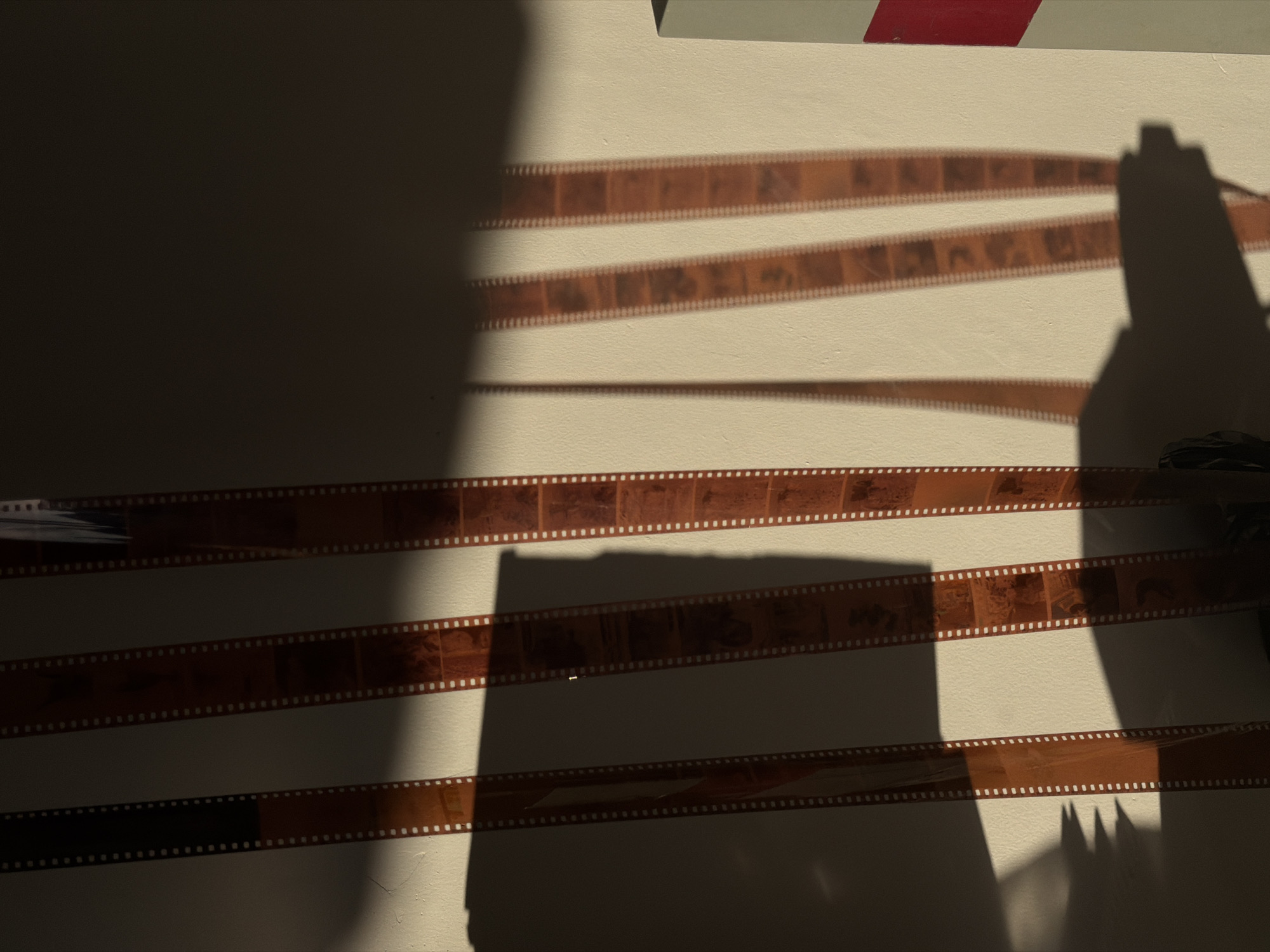A photograph shows several strips of undeveloped film affixed to a white wall. The films are arranged horizontally, with visible perforations along their edges and images visible within each frame. The lighting is stark, casting sharp shadows across the wall, obscuring parts of the film strips. The shadows suggest the presence of an object casting the shadow, possibly a camera, but the object itself is not fully visible in the frame. The photograph appears to be taken indoors. The film strips are undeveloped and have a clear distinction. There are four strips of film. The top of the frame is partially obscured, revealing a narrow section of a gray and red object, likely part of the wall's infrastructure. The photo was taken in North Kuta, Indonesia.