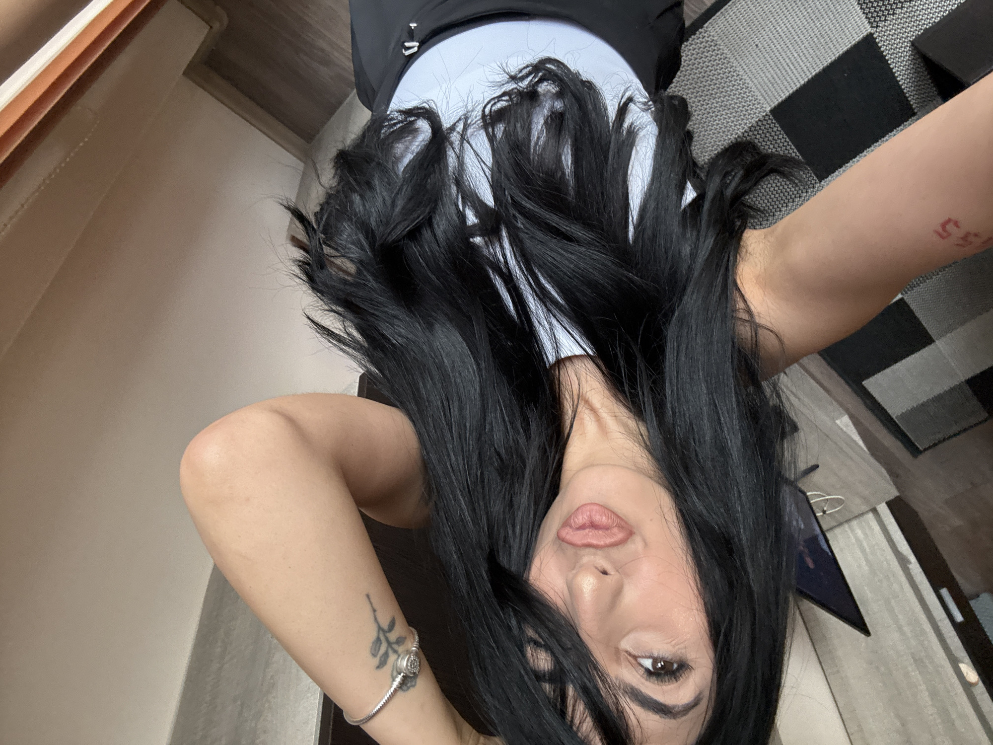 A woman with long black hair is positioned upside down in the frame, with her face centered. She has fair skin, brown eyes, and full lips. She is wearing a light blue tank top with black straps. A silver bracelet is visible on her left wrist, along with a small tattoo of a plant. Another tattoo, consisting of numbers, is seen on her right arm. The background includes a beige wall on the left and a black and gray checkered rug in the upper right corner. A wooden floor and a portion of what appears to be a television and a table are also visible. The exact location in the city of Plovdiv, Bulgaria cannot be determined from the visual data alone.