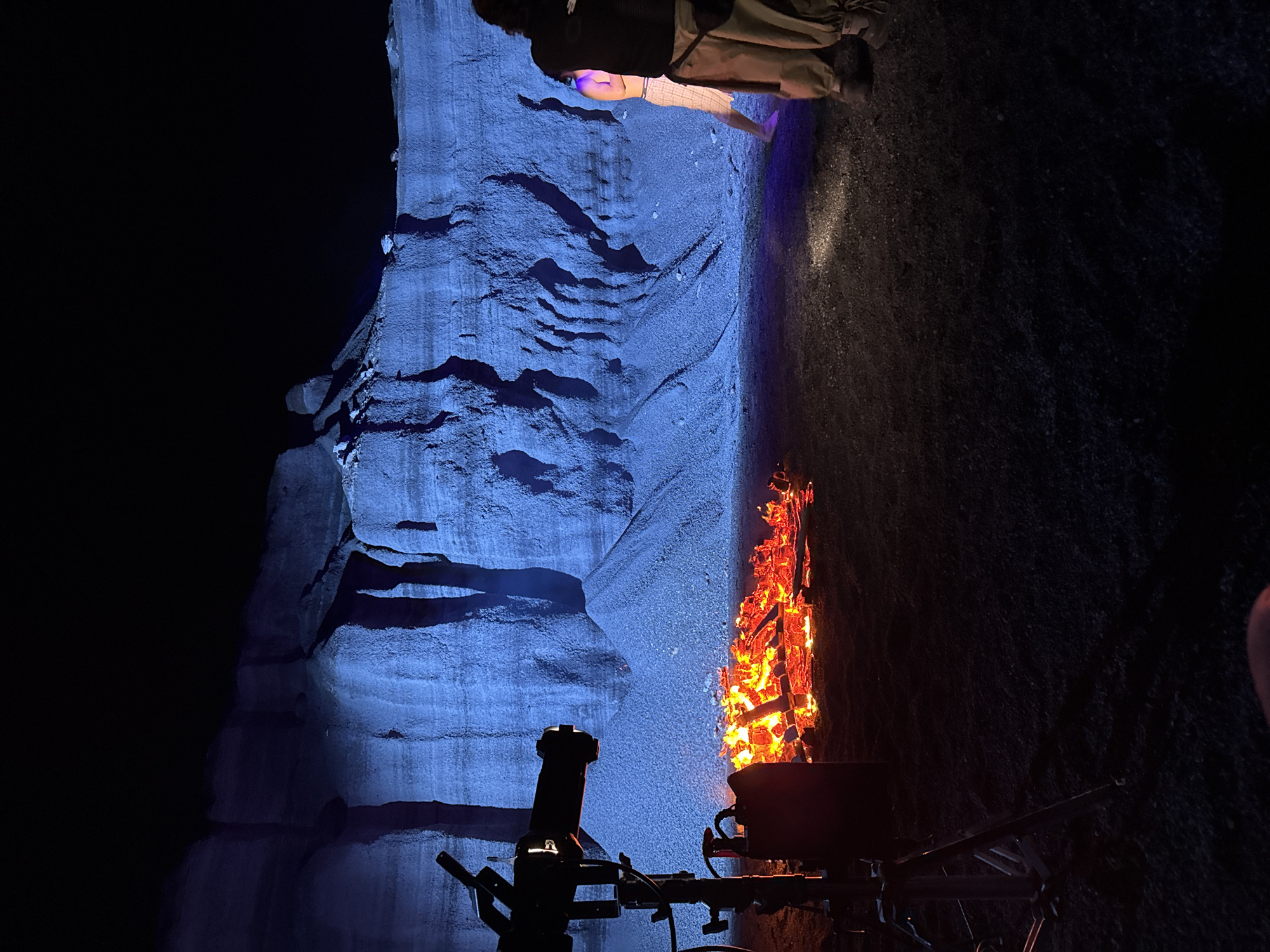 The image shows a dimly lit scene, likely at night, featuring a rock formation illuminated by a blue light on the left. To the right of the rock formation is a contained fire in what appears to be a metal fire pit or container. In the foreground, a black object with a tripod stands, possibly a camera or another piece of equipment. Above the rock formation are two people, seen from the knees down, facing the scene. The ground surface is dark and textured. The location is identified as Teguise, Spain.