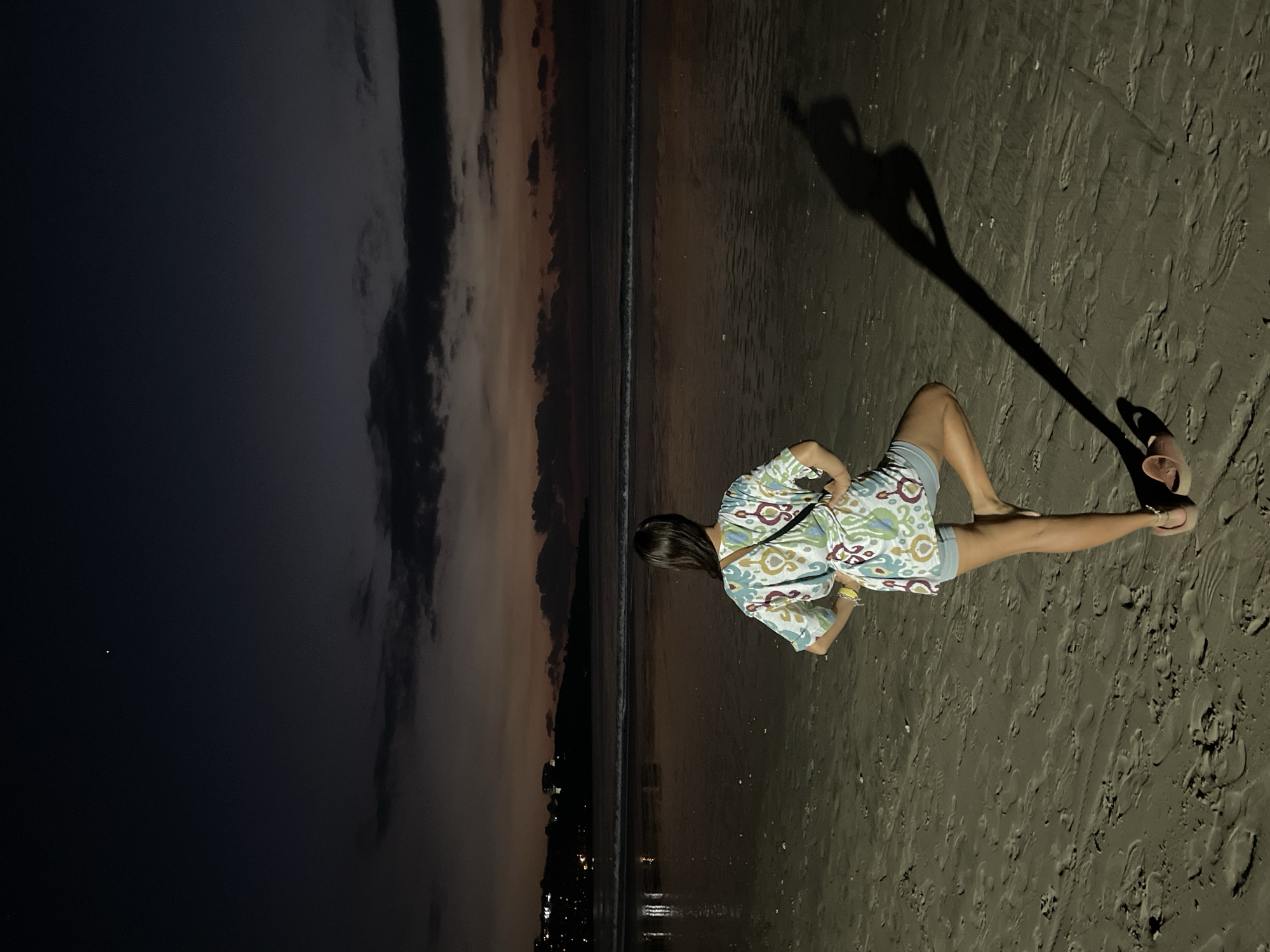 The image depicts a twilight scene on a sandy beach. A person, seen from behind, is the primary subject, positioned towards the right side of the frame. They are wearing a colorful, patterned loose-fitting top and shorts, and appear to be crouching or sitting on the sand, looking out towards the ocean. One flip-flop is visible on the sand near their foot.
The sky in the background transitions from dark, almost black on the left, to hues of orange, pink, and grey around the horizon line, indicative of either dusk or dawn. Gentle waves are breaking along the distant shore. Along the faraway coastline, faint lights are discernible, suggesting a distant town or settlement.
The foreground consists of dark, wet sand, marked with numerous footprints. A very long, distinct shadow of a person, likely the photographer or another individual standing nearby, stretches across the sand from the right side of the frame towards the subject. The overall atmosphere is serene and contemplative. No visible text is present in the image.