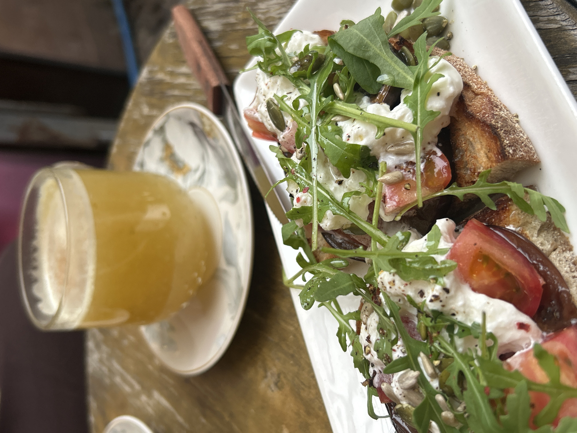 The image shows a table setting, likely in a cafe or restaurant. A rectangular white plate holds slices of bread topped with tomatoes, a white creamy substance (possibly cheese or yogurt), and a garnish of arugula and seeds. A glass of orange juice rests on a saucer to the left of the plate. A knife with a wooden handle is also visible near the plate. The table appears to be made of wood, and the scene suggests an outdoor or semi-outdoor setting. There is no indication of location specific to Saint Petersburg, Russia, in the image.
