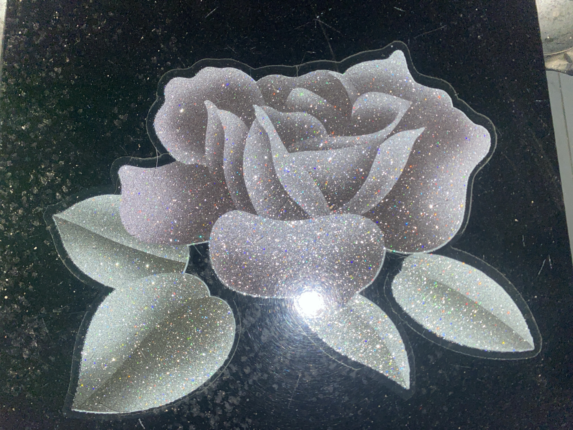 This is a close-up, overhead photograph of a glittery rose design on a black surface. The rose and its leaves are outlined in a thin, dark border and filled with a shimmering silver glitter. The petals of the rose are layered and have a gradient of light and dark silver tones. Some of the glitter pieces appear to be iridescent, reflecting small specks of color. The black surface itself has a speckled texture, possibly resembling granite or a dark, textured paint. There is a bright, circular reflection of light in the lower left quadrant of the rose, obscuring some of the detail. The image does not contain any information about the city of Jauro Yinu, Nigeria.