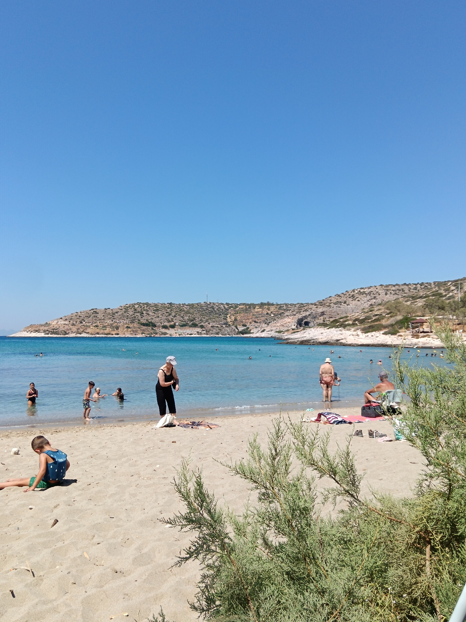 This image depicts a sunny day at Agia Marina beach in Greece. The foreground is dominated by a sandy beach with green foliage in the lower right corner. Several people are visible: a young boy wearing a blue life vest and green shorts is sitting on the sand to the left. In the shallow water near the shore, a group of people are wading and swimming. Further out, a woman in a black swimsuit and a white hat stands on the sand, holding a white bag. In the mid-ground, a man in an orange swimsuit stands in the water, holding a cane, while another man sits on a chair on the sand. The background features a clear blue sea and a hillside covered in sparse vegetation and some buildings. The sky is a bright, cloudless blue.