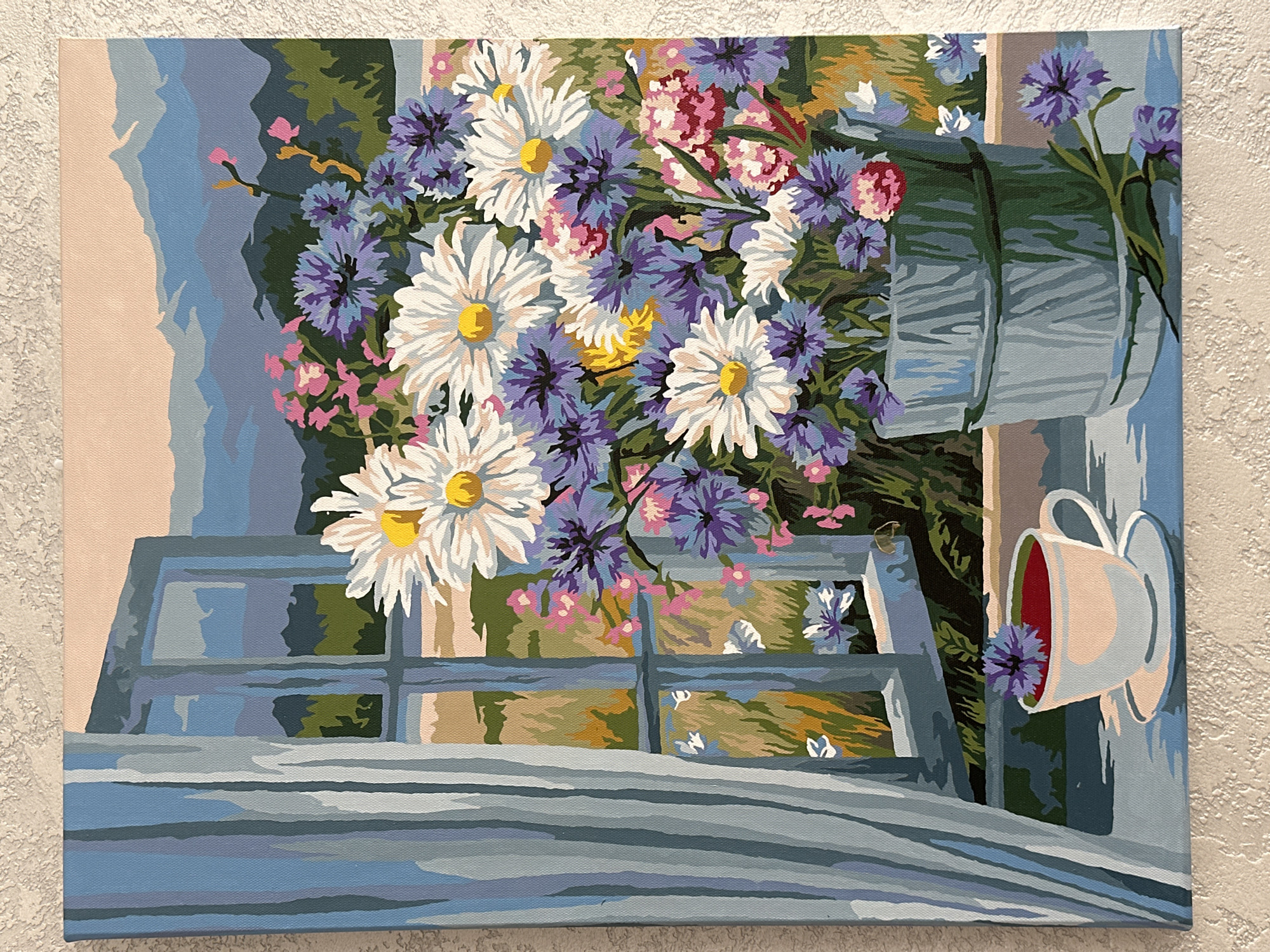 The image depicts a painting of a still life with a floral arrangement and a cup of tea, viewed through a window. The scene is set on a windowsill, with a rustic bucket overflowing with daisies, cornflowers, and pink flowers. To the left, a sheer blue curtain hangs. A white teacup filled with a red liquid, possibly tea, sits on the windowsill, adorned with a single cornflower. Through the open window, a landscape is visible, featuring rolling hills and distant mountains under a soft, peachy sky. The style of painting is reminiscent of a "paint by numbers" artwork, with bold blocks of color and clear outlines. There is no indication of the city of Yessentuki, Russia in the image.