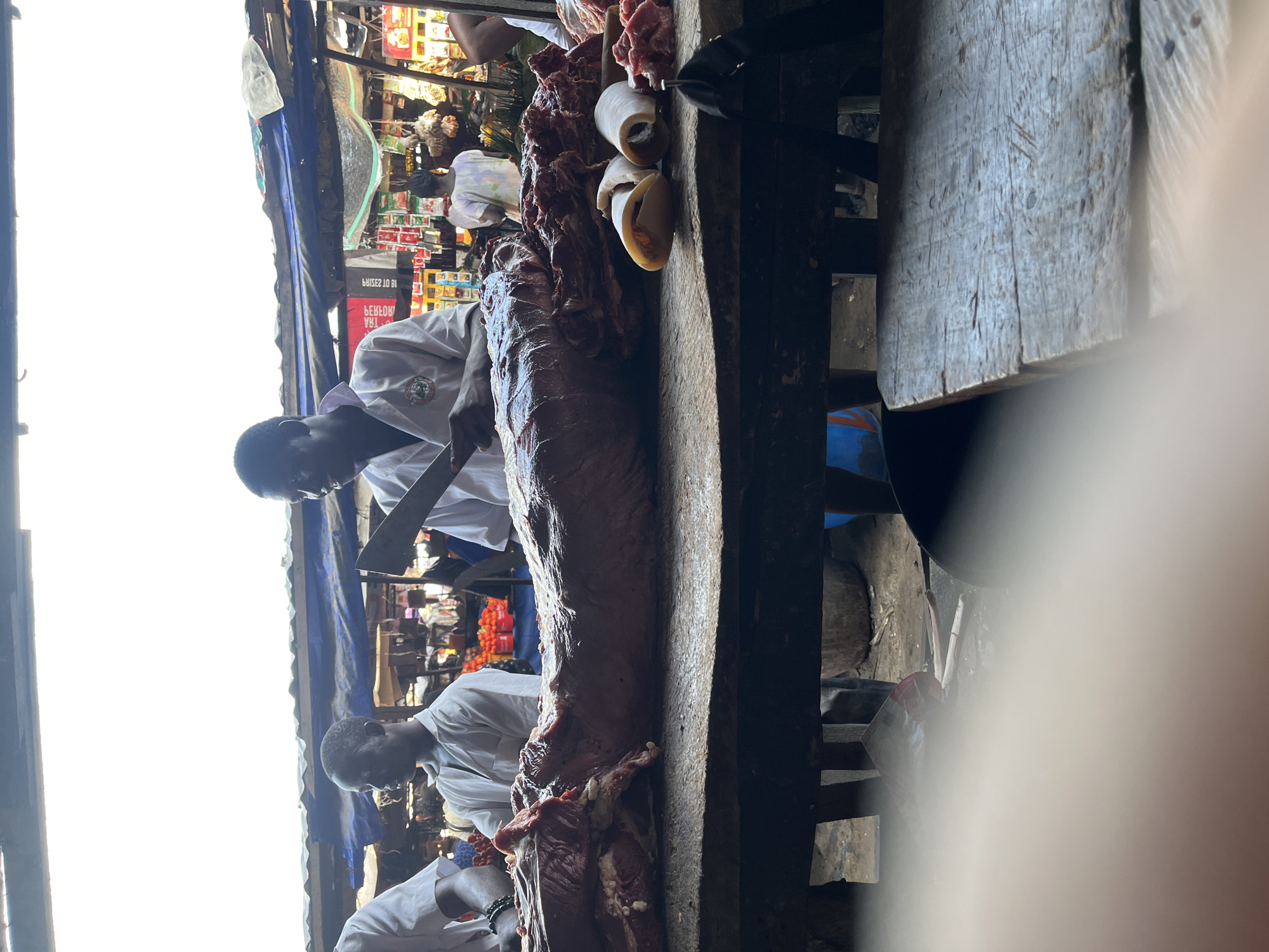 In a bustling open-air market in Ado, Nigeria, two men wearing white shirts are butchering a large cut of meat on a wooden cutting board. One man is actively using a cleaver to cut the meat. Behind them, a variety of stalls selling goods, including produce, are visible. The scene is illuminated by bright daylight. The overall environment appears to be a typical, busy marketplace.
