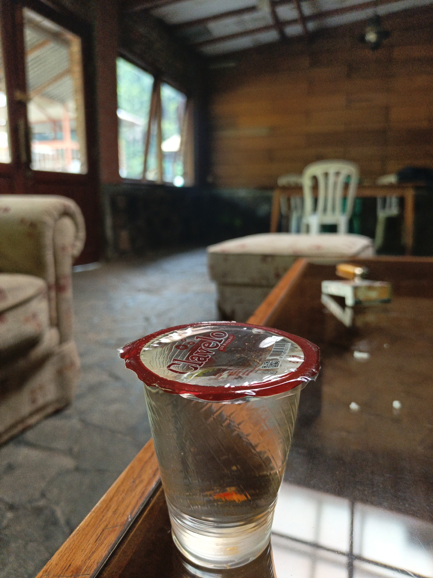 The image depicts an indoor scene with a focus on a plastic cup sealed with a red foil lid, resting on a wooden table. The cup appears to contain a dark substance and possibly some liquid. The table surface is reflective, showing a faint reflection of a grid-like pattern, possibly from a window or light fixture. In the background, a floral-patterned armchair is visible to the left, and beyond that, a room with wooden walls and what appear to be windows with a view of greenery. A white chair and a wooden table are also present in the mid-ground. The overall environment suggests a rustic or casual setting, possibly a dining area or lounge. No people are visible in the image.