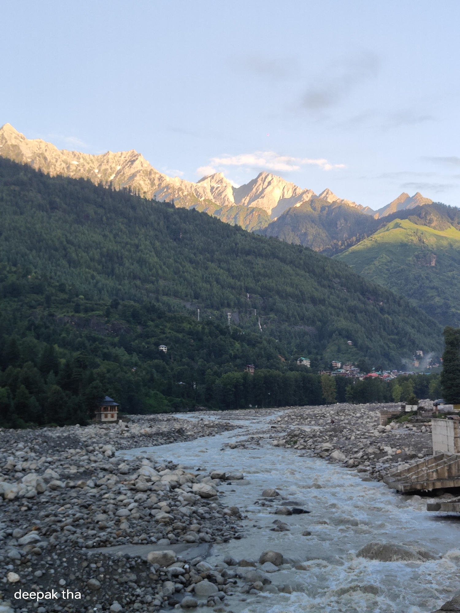 A landscape photograph depicts the Beas River flowing through a valley in Vashisht, India, with steep, tree-covered mountains in the background. The mountains are topped with snow-capped peaks illuminated by sunlight. A small, multi-story structure with a dark roof and light-colored walls sits on the riverbank. The river is wide and appears to be flowing rapidly, with many large, smooth stones visible along its bed and banks. The sky is a pale blue with a few scattered clouds. The overall impression is one of a serene and natural environment.