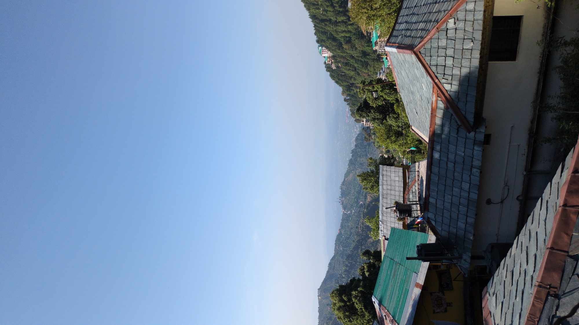 The image is an angled outdoor shot capturing rooftops, a hillside, and a clear blue sky. The scene suggests a location in the mountains, possibly the city of Haini, India, given the landscape characteristics.
The foreground features an array of rooftops. The roofs exhibit varied designs, some are of blue-grey slate tiles with reddish-brown edges. Buildings appear to be constructed close to each other, with an exterior wall and window visible along one side. The roofs indicate varying levels of wear and construction materials.
In the middle ground, a mountain slope covered with lush green trees occupies the central horizontal section of the frame. The topography is steep and the forest vegetation is dense. The distant hillside merges into a light, hazy atmosphere, which reduces color saturation and detail.
The background is dominated by a clear, bright blue sky. There are no visible clouds or other atmospheric phenomena, indicating clear weather conditions.