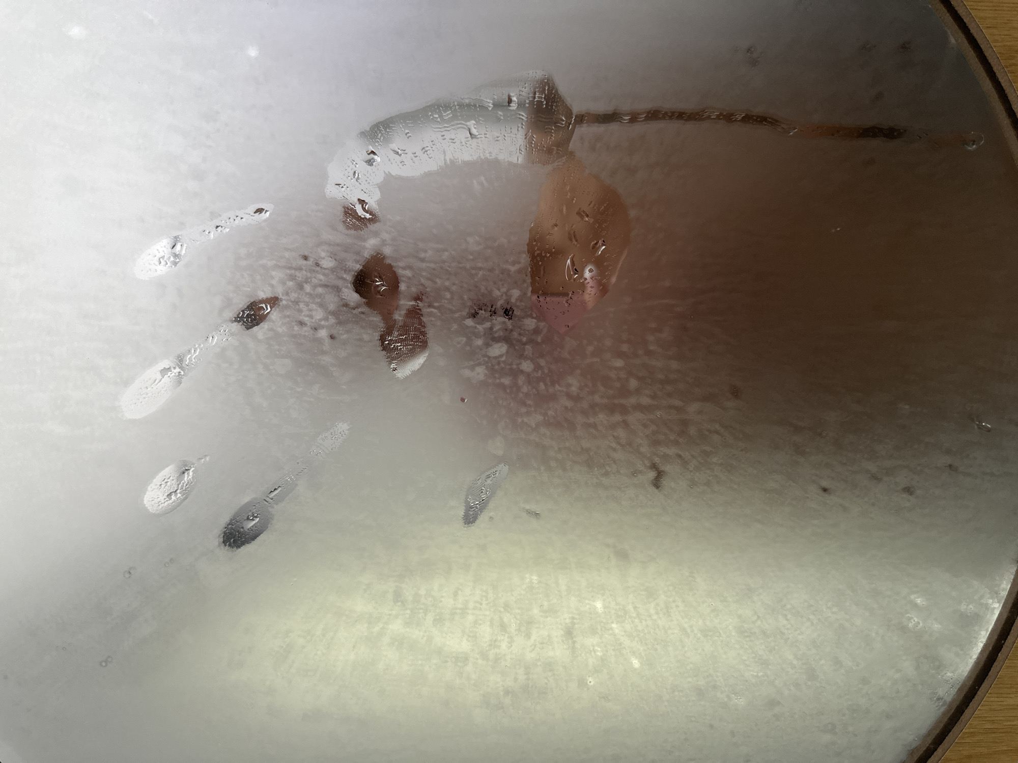 The scene is described as being in Mũi Né, Vietnam. The image displays a clear impression of a human left hand, palm facing the viewer, on a severely fogged or steamed transparent surface, likely glass or a mirror. The handprint, positioned centrally, has cleared away the condensation, revealing blurred, indistinct forms and skin tones (light brown, reddish-pink) behind the surface, suggesting the presence of a person. Distinct water droplets of various sizes adhere to the surface, particularly concentrated around the edges of the handprint and the individual finger impressions. A significant streak of accumulated water runs vertically down the lower right portion of the surface. The background of the unfogged areas is uniformly bright and diffuse, indicating soft, even illumination. The lower edge of the circular surface is framed by a thin, dark wooden border.