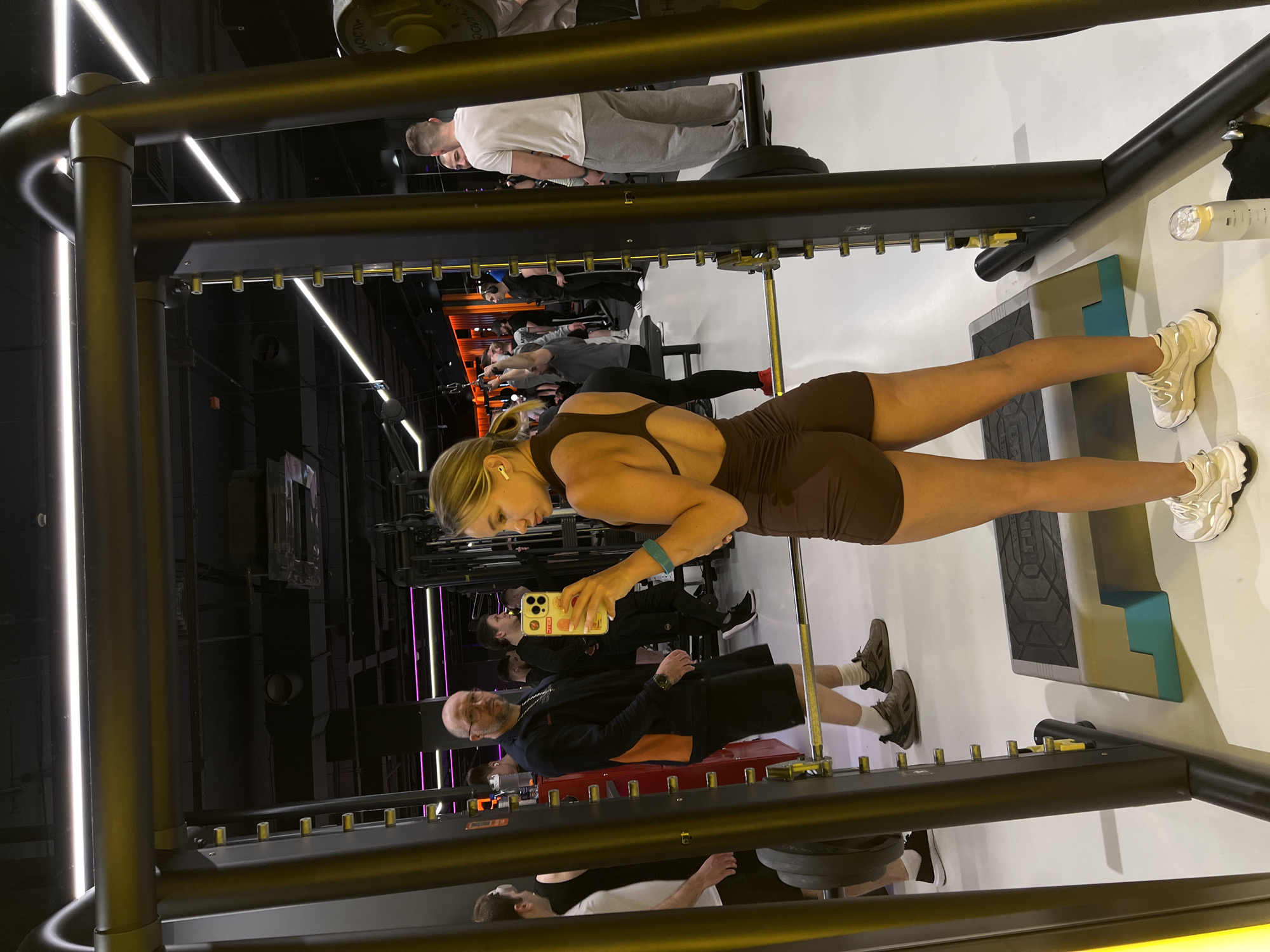 The image, rotated clockwise, depicts an indoor scene within a modern fitness center or gym. The main subject is a blonde woman in the foreground, taking a mirror selfie using her smartphone. She wears a dark brown athletic set consisting of a top with a cut-out back and matching shorts. She also has white chunky sneakers and white earbuds, with a blue fitness band on her left wrist.
The woman is positioned in front of a reflective surface, which shows various other gym patrons and equipment in the background. Visible in the reflections are several individuals engaged in exercise. An overhead reflection shows a man in a white t-shirt and grey sweatpants near what appears to be weightlifting equipment. Another man, wearing a black long-sleeved shirt and black shorts, is visible in a lower reflection.
The gym features robust black metal structures, possibly part of a squat rack or Smith machine, with rows of golden pins for height adjustment. The facility is illuminated by bright linear LED lights, with both white and purple lights creating a dynamic ambiance against dark walls. A grey and teal step platform is on the floor in front of the woman. The overall scene suggests an active and contemporary gym environment in Voronezh, Russia, with people focused on their workouts. No distinct text is clearly legible on any objects or clothing.