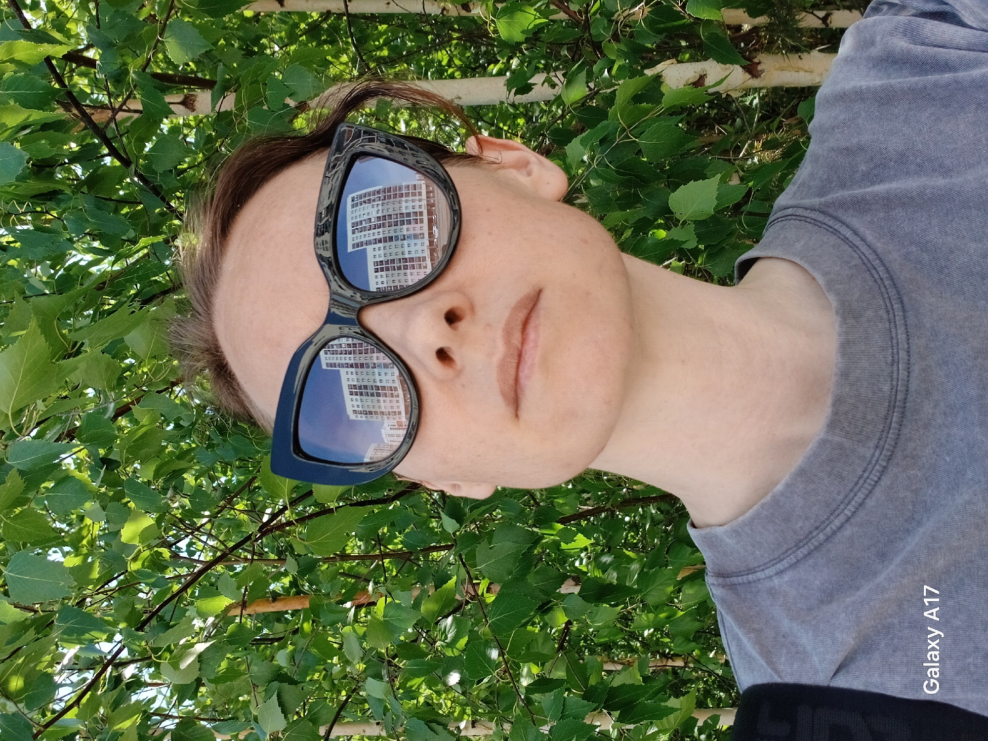 A woman is facing the camera with a building reflected in her sunglasses. She is wearing a gray shirt, and green foliage is behind her. The building reflected in her sunglasses appears to be a multi-story residential building, likely a typical Soviet-era apartment block, possibly located in a residential area of Yekaterinburg, Russia. The text "Galaxy A17" is visible on the right side of the image.