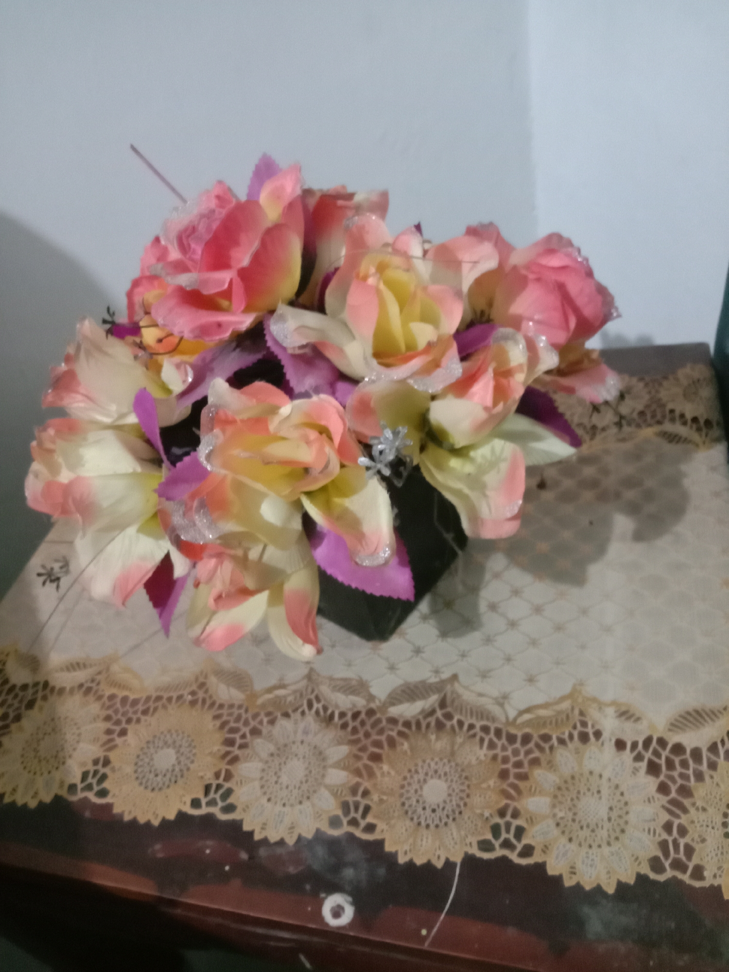 A bouquet of artificial flowers, primarily in shades of pink, yellow, and cream with glitter accents, is arranged in a dark square vase. The vase sits on a table covered with a light-colored, patterned tablecloth. Below the tablecloth, a dark wooden surface is visible, possibly the tabletop of a desk or table. A decorative lace trim with floral motifs runs along the edge of the tablecloth. The background consists of a plain light blue wall. The scene is lit by ambient light, casting shadows from the flowers and vase onto the tablecloth and the wall. The image quality is low, with a blurry appearance. The location is identified as Kwandang, Indonesia, implying this is a domestic setting. There are no people or animals present in the image, and no actions or interactions are depicted.