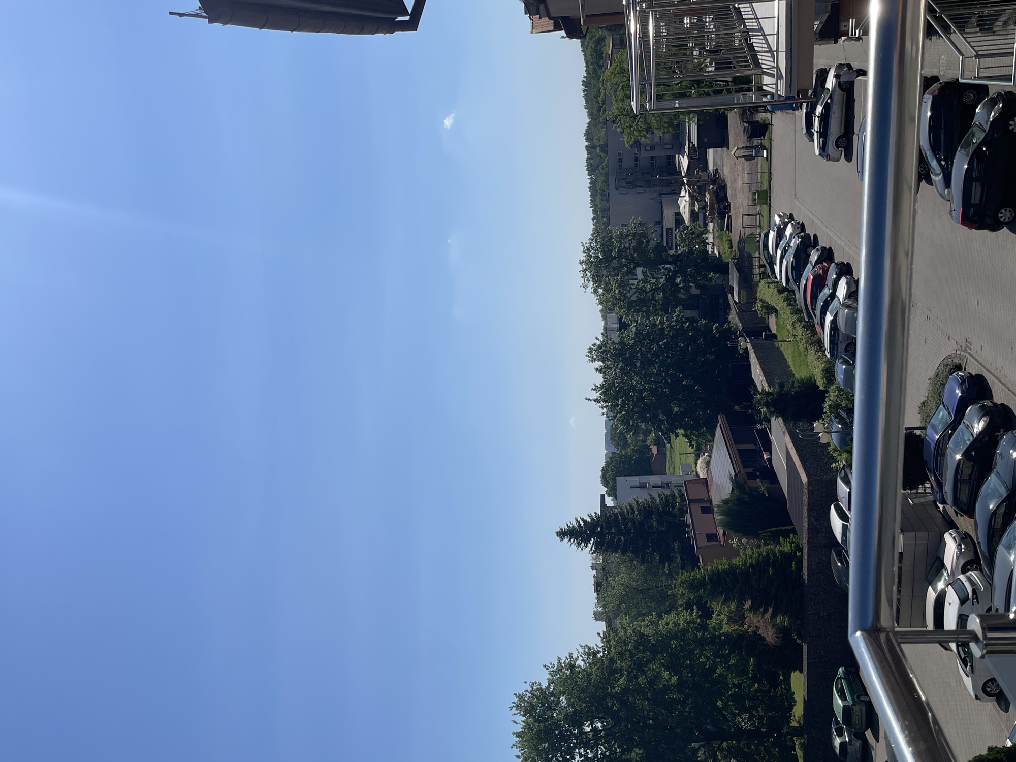 View from a balcony overlooking a parking lot with multiple cars parked next to buildings and green spaces under a clear blue sky. The location is in an urban area. A shiny metal railing of the balcony is visible in the foreground.