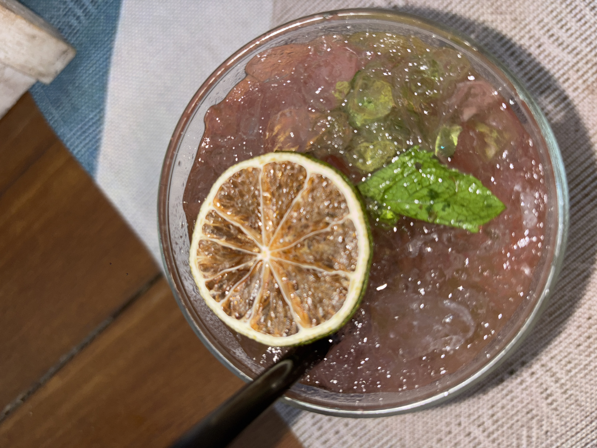 This is a top-down, close-up shot of a glass of pinkish-red liquid, likely a beverage, filled with ice. A dried slice of lime is placed on top of the drink, positioned towards the left side of the glass. To the right of the lime slice, a sprig of mint leaves adds a touch of green to the composition. The drink appears to have a slightly gelatinous or slushed consistency, with some translucent green chunks visible throughout. The glass is clear and sits on a textured, light-colored surface, possibly a tablecloth or placemat. A dark-colored straw is partially visible entering the glass from the bottom left. In the background, out of focus, a wooden surface and a striped blue and white fabric are present, suggesting an outdoor or casual setting. The lighting indicates it might be daytime, with some reflections and highlights on the drink's surface. There is no text visible in the image.