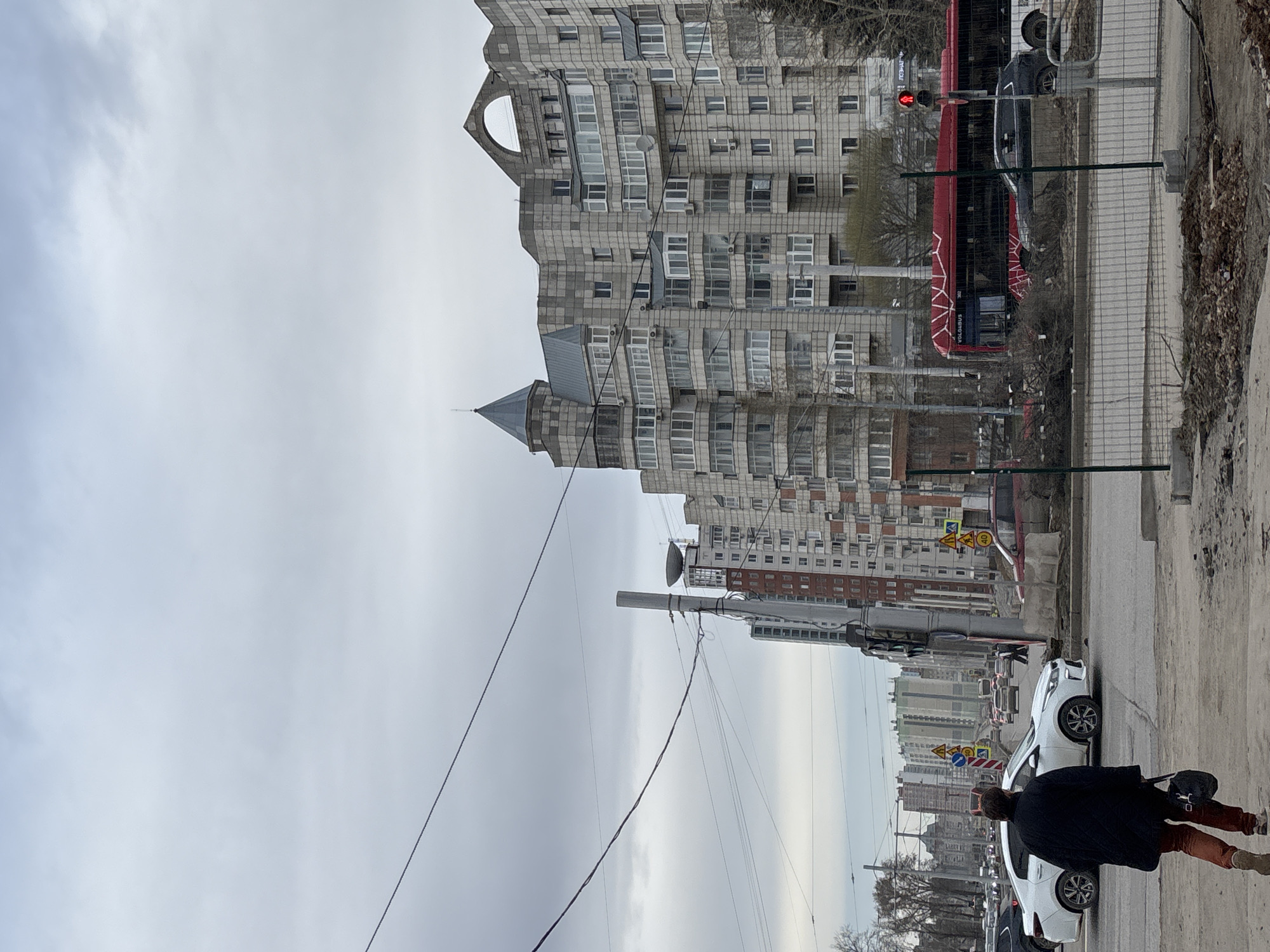 The image shows a street scene in Perm, Russia. In the foreground, a person wearing a dark coat, reddish-brown pants, and light-colored boots is walking along the sidewalk. The sidewalk is adjacent to a street with parked and moving cars, including white compact vehicles. A red and white bus is stopped at a traffic light, which is showing a red signal. In the background, there are several tall residential buildings of Soviet-era architecture, primarily constructed of gray concrete with numerous windows and balconies. An overhead network of utility poles and power lines are present. The sky is overcast with gray clouds. There are construction barriers or fencing along parts of the street.