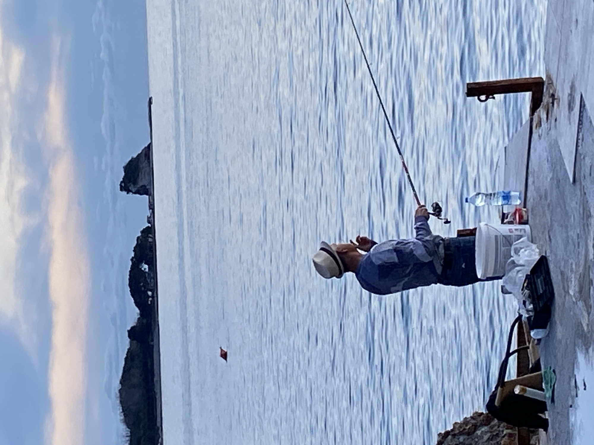 An outdoor image shows a man fishing on a concrete pier next to blue water. The man wears a light-colored hat and a blue shirt. He holds a fishing rod. A water bottle and other items are placed next to him on the pier. The pier is connected to a rocky area. Across the water is an island with green trees and a rocky cliff. A small, square, red object floats in the water. The location is likely near the coast of Budva, Montenegro, based on the landscape features.