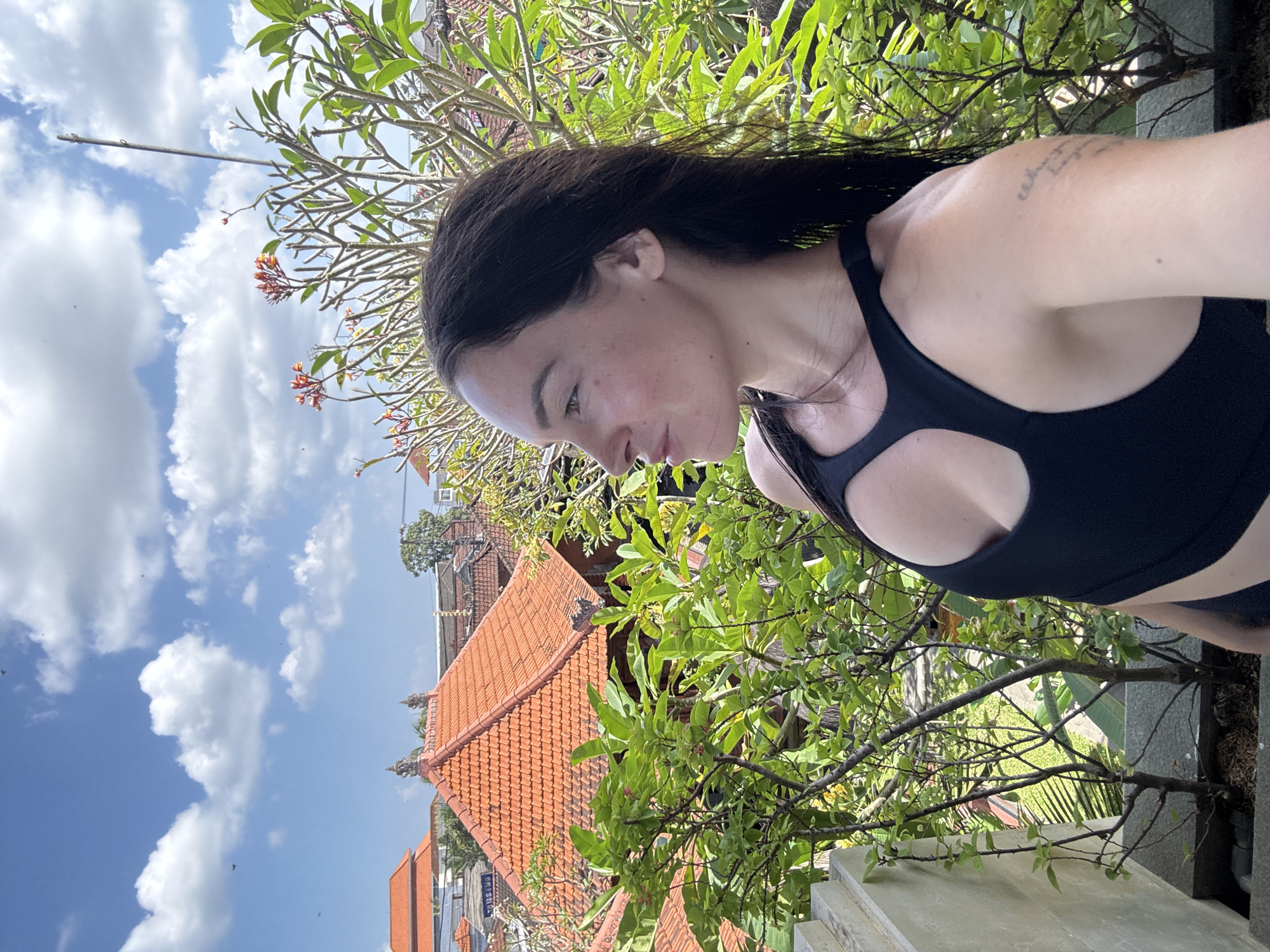 A woman with long dark hair is pictured outdoors, looking to her left in profile. She is wearing a black sports bra. Behind her, there are green trees and shrubs with orange flowers, and in the background, there are buildings with orange tiled roofs. The sky is blue with white clouds. The scene appears to be in North Kuta, Indonesia, likely a rooftop or balcony setting.