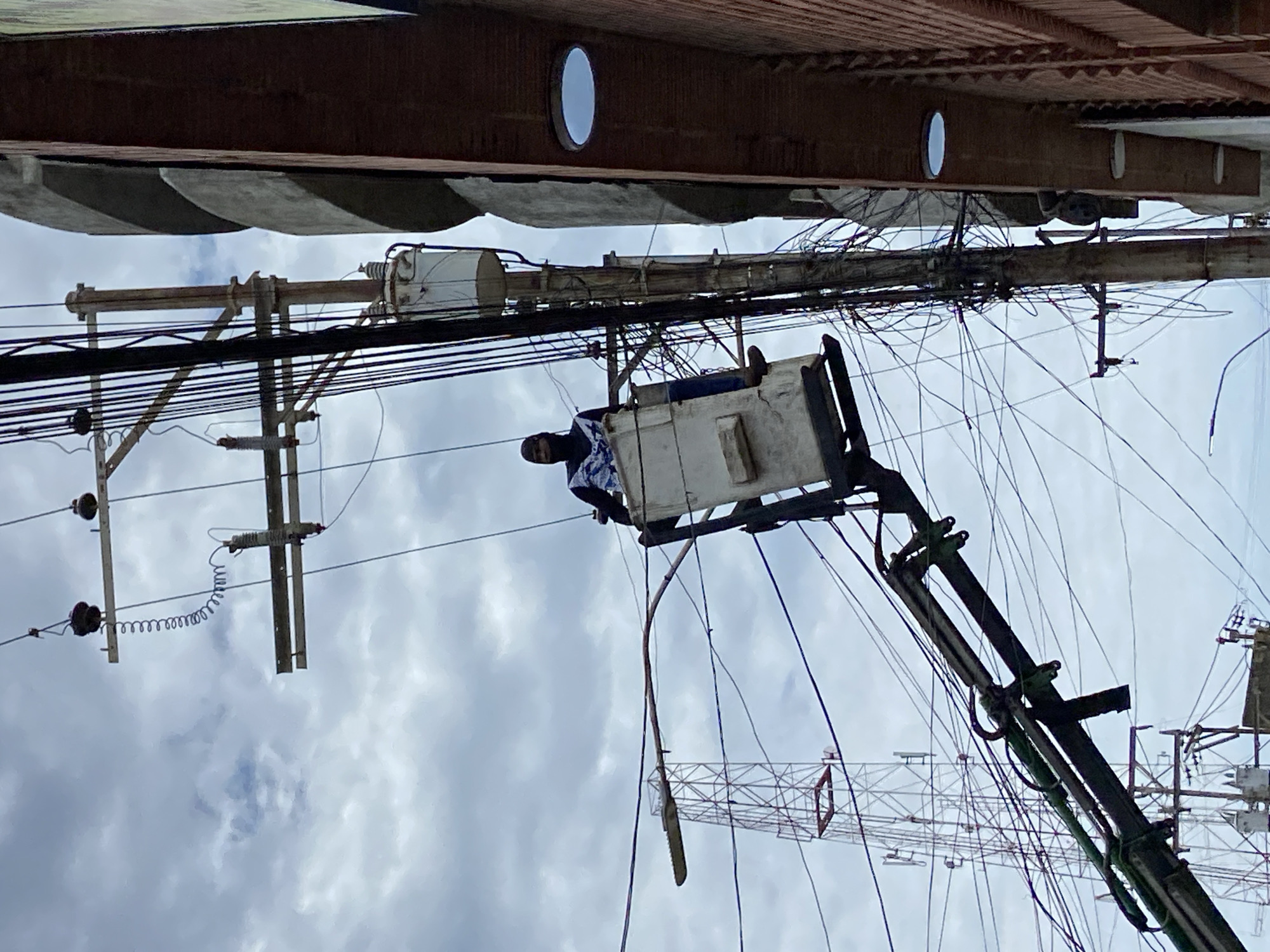 The image depicts a man in a cherry picker working on power lines in Higuerote, Venezuela. The cherry picker is extended upwards towards a dense network of electrical wires and utility poles against a cloudy sky. The man appears to be wearing a dark shirt and possibly a head covering. Overhead, there is a building with rounded windows. The background includes other utility poles and lines.