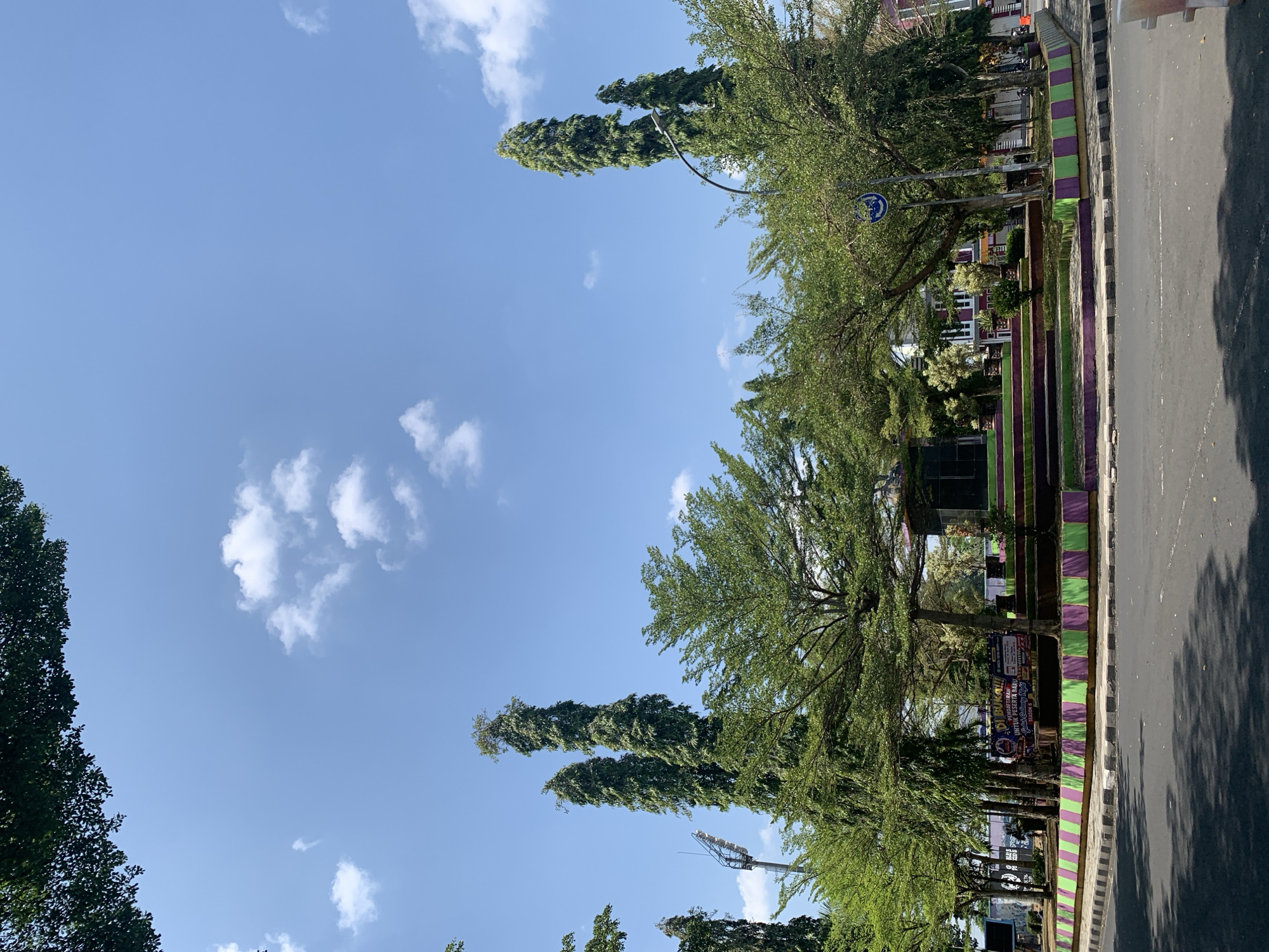 This image captures a street-level view of a roundabout in Ciamis, Indonesia, likely in the city center given the presence of a monument and surrounding buildings. The sky is a clear, bright blue with scattered white clouds, indicating a sunny day. Dominating the scene are several tall, slender trees with lush green foliage, some reaching significant heights and appearing to be cypress trees. These trees are interspersed with shorter, bushier trees that form a dense green canopy around a central monument.
The monument itself is built on a tiered, stepped structure, with the steps painted in alternating shades of purple and green. The base of the roundabout is also decorated with the same purple and green checkered pattern. A signpost with a blue circular traffic sign is visible near one of the taller trees. Behind the roundabout, a few buildings are partially visible, suggesting an urban environment. One building has a traditional red tile roof, while others appear to be more modern. A banner with Indonesian text is displayed near the monument, possibly advertising an event or service. The road in the foreground is asphalt, with clear lane markings and a raised curb. The overall impression is a well-maintained public space with ample greenery, typical of a city park or a prominent intersection in Indonesia.
