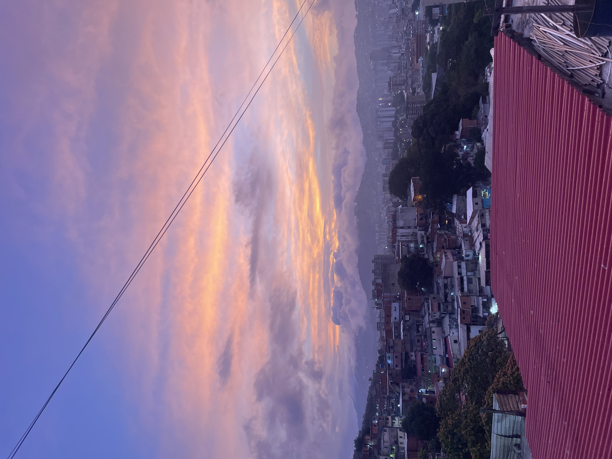 The image shows a high-angle view of Caracas, Venezuela, at dusk or dawn. The sky is filled with layered clouds colored with shades of orange, pink, and purple. In the foreground, a red corrugated metal roof is visible, likely part of the building from which the photo was taken. Power lines stretch across the sky. Below the sky, a densely packed urban landscape is visible, composed of buildings and houses stretching up a hillside. Some buildings have lights on. In the distance, mountains are visible, partially obscured by haze or clouds.