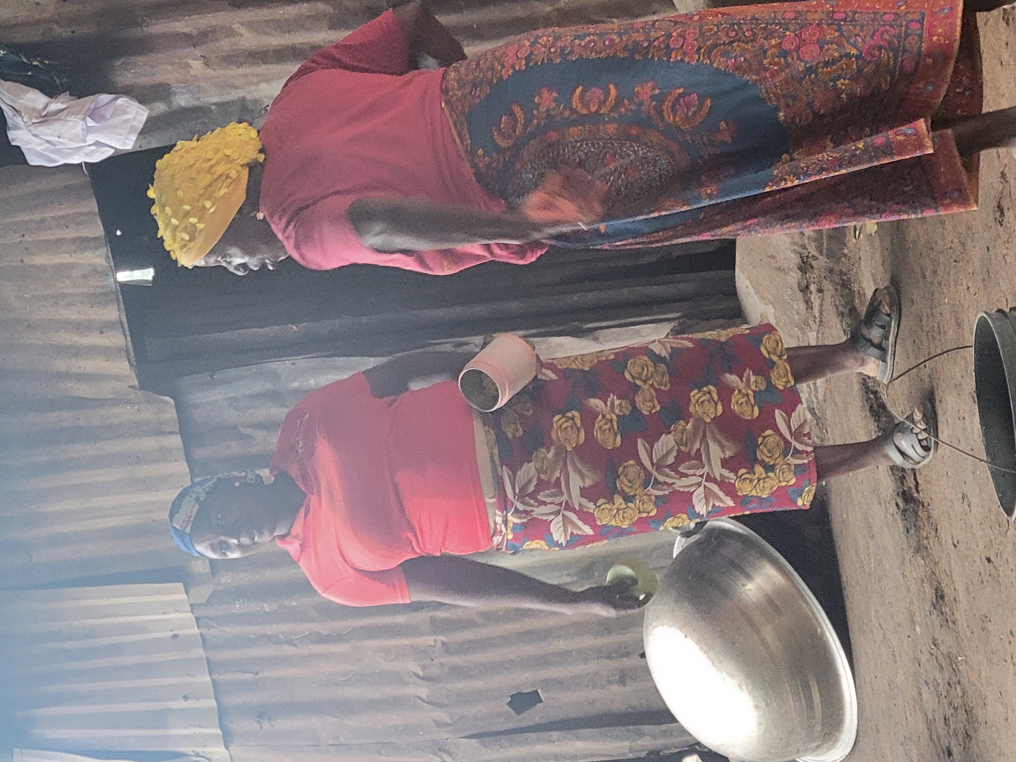 Two women are depicted standing in what appears to be a modest indoor or semi-outdoor setting, possibly in Madalla, Nigeria. The image is oriented vertically, showing the women and their surroundings from a slightly elevated perspective.
The woman in the foreground wears a short-sleeved red top, a long maroon skirt adorned with yellow and white floral patterns, and a blue head covering. She has dark skin and is wearing open-toed sandals. Her left arm is extended, holding a light green, possibly spherical, object over a large, shiny metal bowl placed on the earthy ground. A light pink cup is visible near her waist, tucked into her skirt or held.
The second woman stands further back, to the right of the first. She also wears a short-sleeved red top, paired with a long, dark blue skirt featuring intricate red and gold patterned designs. Her head is covered with a vibrant yellow, textured headwrap. She is standing with her hands down, looking towards the left side of the frame.
The background and walls of the structure appear to be made of corrugated metal sheets, showing some signs of wear and discoloration. The ground is rough and appears to be packed earth or dirt. A crumpled white cloth is visible in the upper left background. The overall scene suggests a domestic environment, possibly engaged in daily chores like food preparation. There is no visible text in the image.