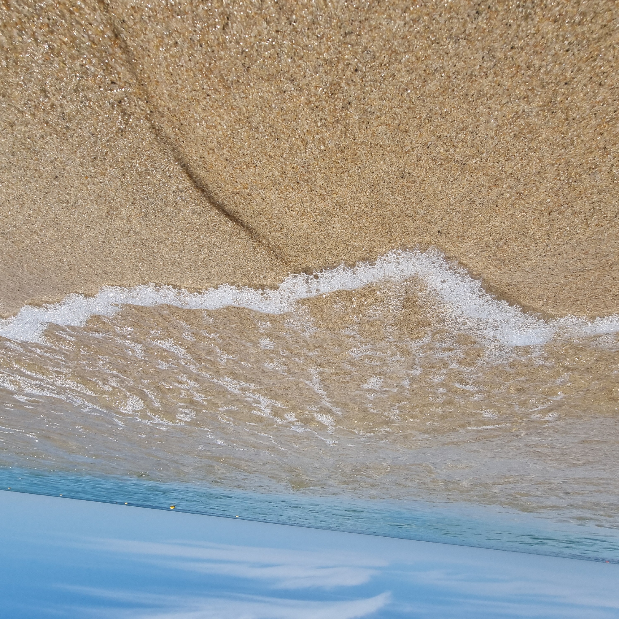 The image captures a low-angle perspective of a sandy beach with gentle waves lapping at the shore under a clear blue sky with scattered white clouds. The foreground is dominated by the wet, golden sand, with a thin line of white foam from the incoming wave tracing a curve across the beach. In the midground, the shallow water is transparent, revealing the sandy seabed beneath. The horizon line is clearly visible, separating the ocean from the sky. A few yellow buoys are sparsely dotted in the ocean, indicating a designated swimming area or marker. The scene suggests a tranquil day at the beach, likely in the city of Gangneung-si, South Korea, known for its beautiful coastline. There are no people or animals visible in the frame, emphasizing the natural environment.