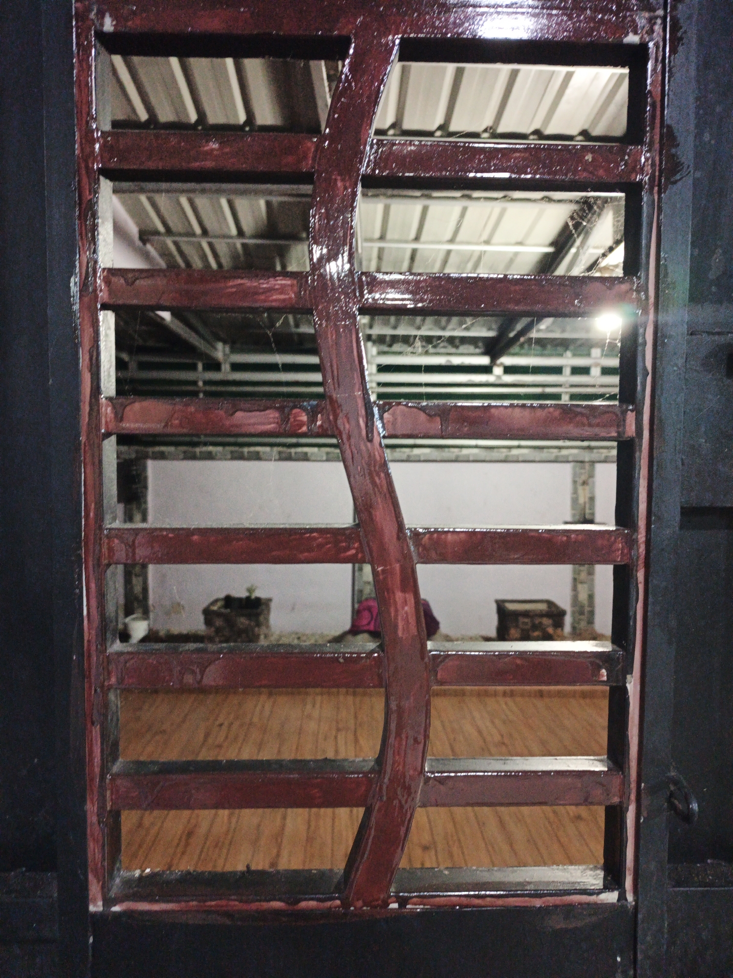 This is a medium shot, eye-level, indoor photograph taken at night. The foreground features a dark, weathered, red-painted metal gate with a vertical, wavy decorative element in the center. Through the gate's horizontal slats, we see an interior space with a light-colored, corrugated metal ceiling supported by metal beams, suggesting a workshop or storage area. Below the ceiling, a wooden floor with a visible grain pattern is present. In the middle ground, on the wooden floor, are several objects: a small, dark planter with a seedling, a white bowl, and a small, dark rectangular box. The lighting appears to be artificial and dim, with a bright light source visible in the upper right quadrant of the image, creating a lens flare. The overall impression is one of disuse or neglect, with dust or cobwebs visible in some areas. The location is identified as Kelapapati, Indonesia.