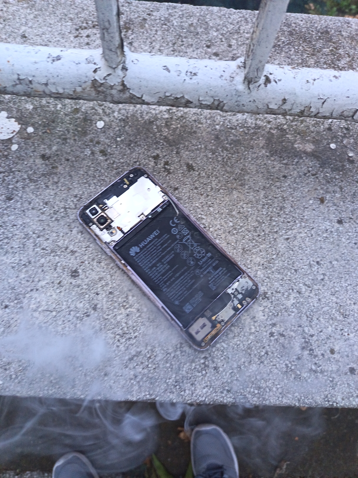 A damaged Huawei smartphone is lying on a concrete surface next to a metal railing. The back of the phone is open, exposing the battery and internal components. Smoke is visible rising from the concrete surface near the phone. Two feet wearing grey sneakers are visible at the bottom of the frame, suggesting a person is standing nearby. The environment appears to be outdoors, possibly in an urban setting. The image was taken in the city of Yambol, Bulgaria.