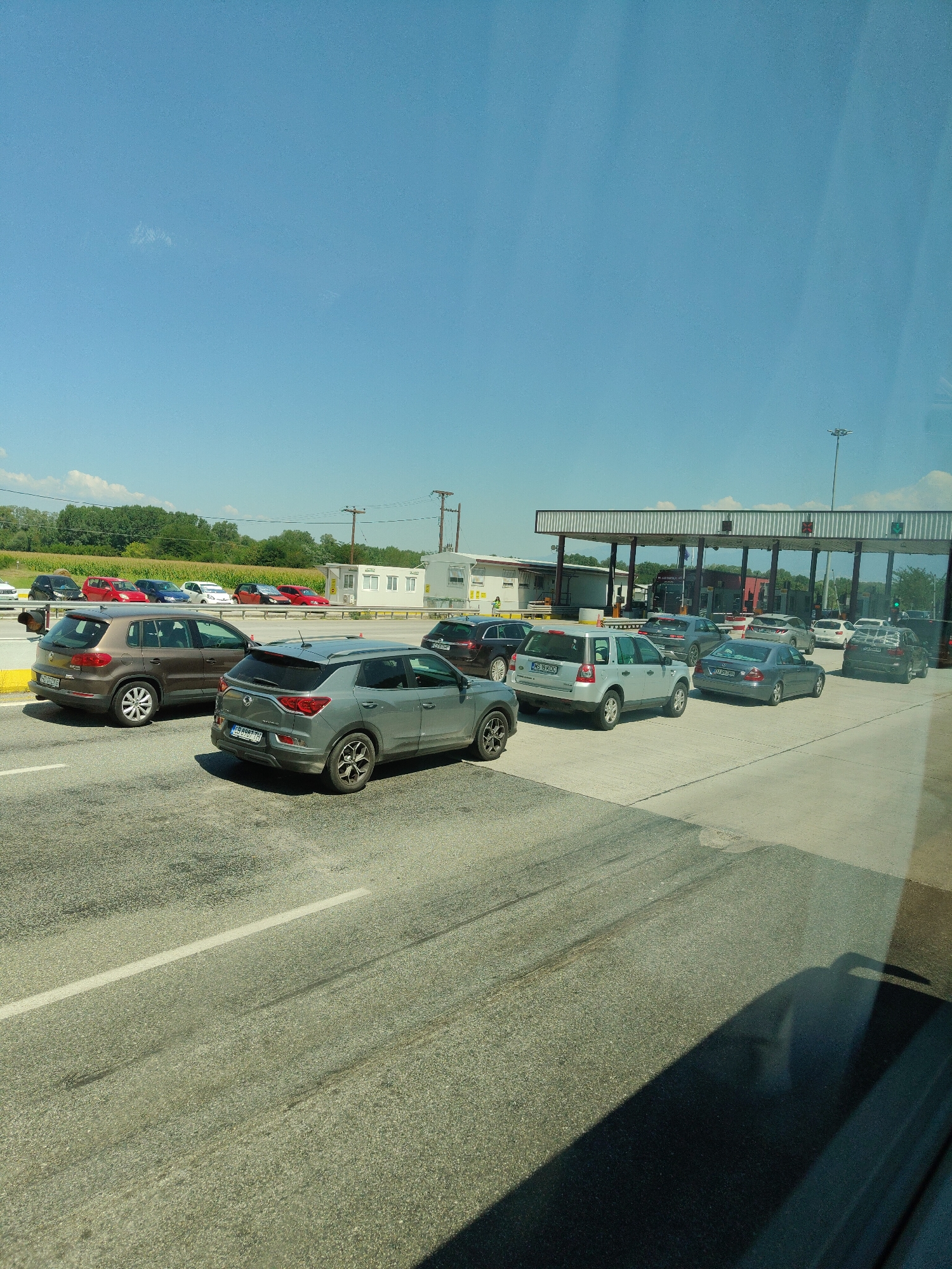 A convoy of vehicles is queuing at a toll booth plaza under a clear blue sky. The scene appears to be set at a border crossing or a highway toll collection point. In the foreground, a gray SUV is parked, followed by a brown SUV. Further back, several other cars are lined up in multiple lanes, including a silver sedan and a dark-colored SUV. The toll booths themselves are visible in the mid-ground, with a white building and various signs. The environment is paved and appears to be outdoors, with trees and fields visible in the background. The image suggests a pause in traffic flow due to the toll collection process. This location is identified as being in the Municipal Unit of Promachonas, Greece.