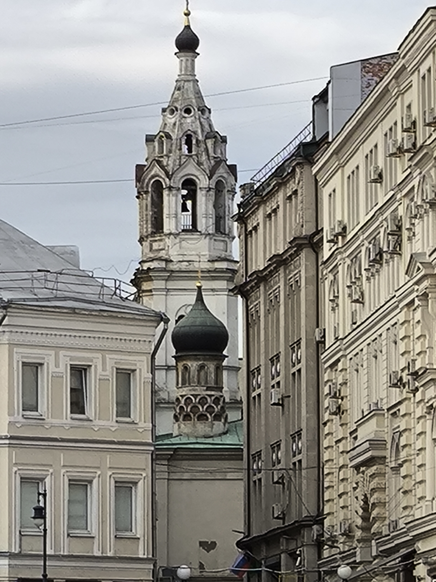 The image depicts a street scene in an urban environment, likely in a historic city. The focal point is a tall, ornate church tower with a steeple and a black dome-shaped structure. The tower appears to be white or light-colored and features arched openings and decorative elements. Several buildings surround the church tower, with varying architectural styles. One building has a light facade with multiple windows, while another has a darker, more austere appearance. Several air conditioning units are attached to one of the buildings. The sky is overcast, creating a soft, diffused light. The overall scene suggests a densely populated area with a mix of religious and secular architecture. The location is Unknown, Unknown.