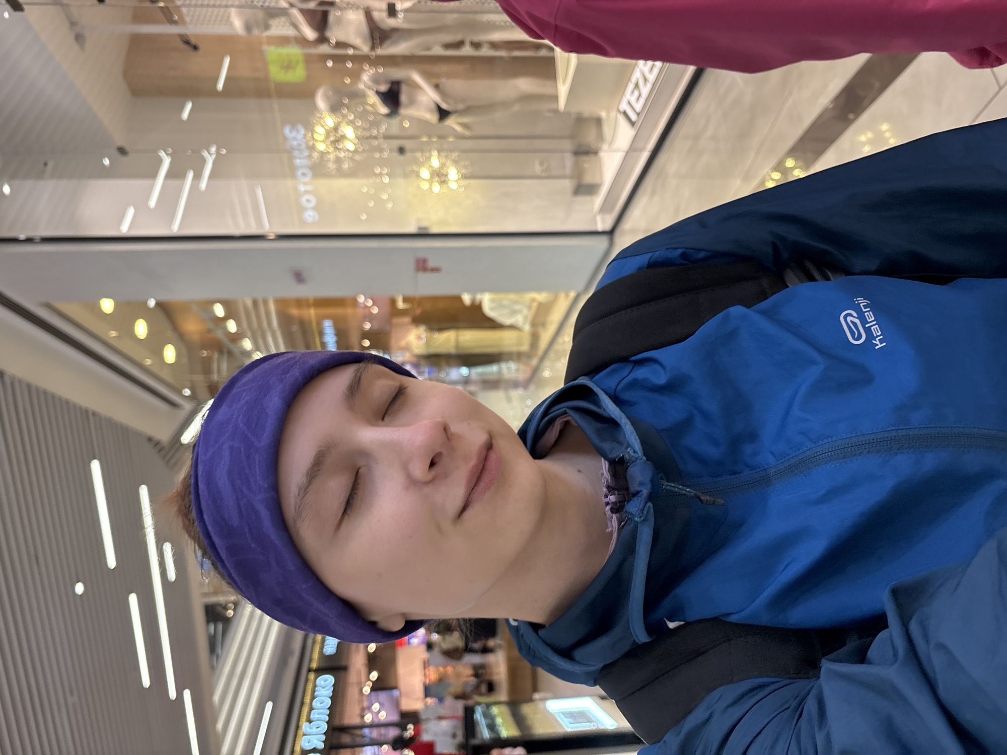 The image shows a person wearing a blue jacket and a purple headband in what appears to be a shopping mall in Perm, Russia. The person has their eyes closed and seems to be in a relaxed state. In the background, there are store fronts and interior lighting, suggesting a retail environment. The word "Яблоко" is visible on one of the storefronts. Other people are also visible, adding to the mall's crowded atmosphere. The view is tilted slightly, giving a sense of the perspective from which the photo was taken.