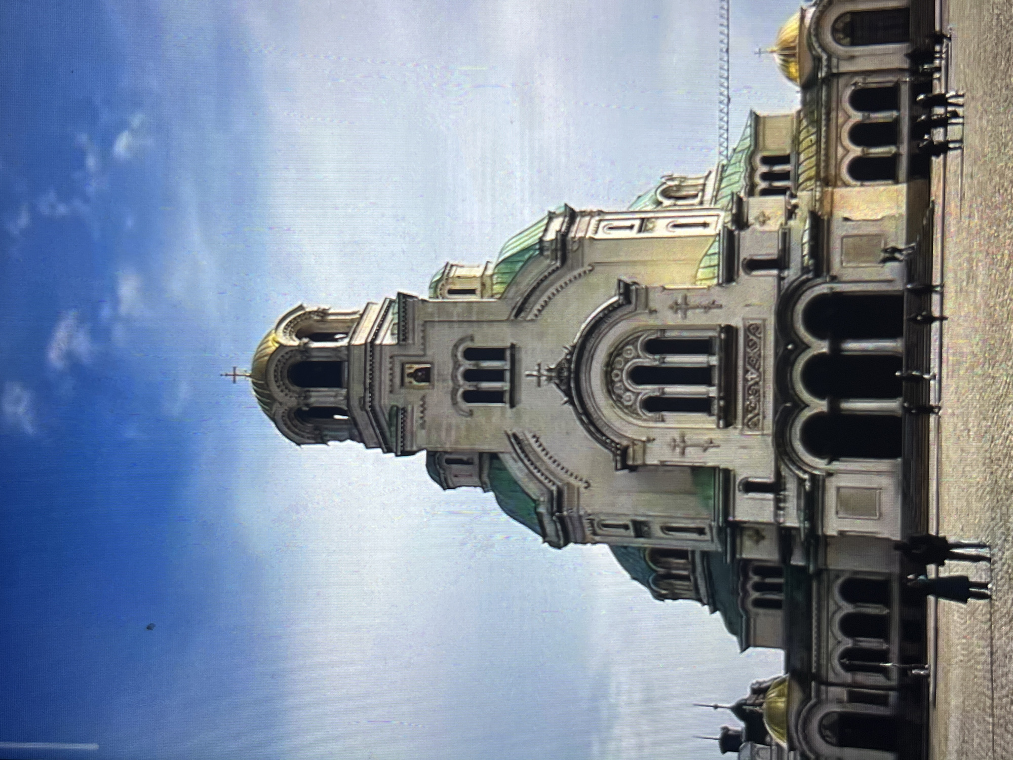 The image shows a large, ornate cathedral with multiple golden domes against a blue sky with scattered clouds. The cathedral is primarily white or light-colored, with green accents on the roofs of some of the domes. The architecture is detailed, featuring arches, decorative elements, and crosses. Several people are standing or walking along the side of the cathedral. The ground appears to be paved with stone. There is no indication that the location is in Tinaquillo, Venezuela. The building appears to be the Alexander Nevsky Cathedral in Sofia, Bulgaria.