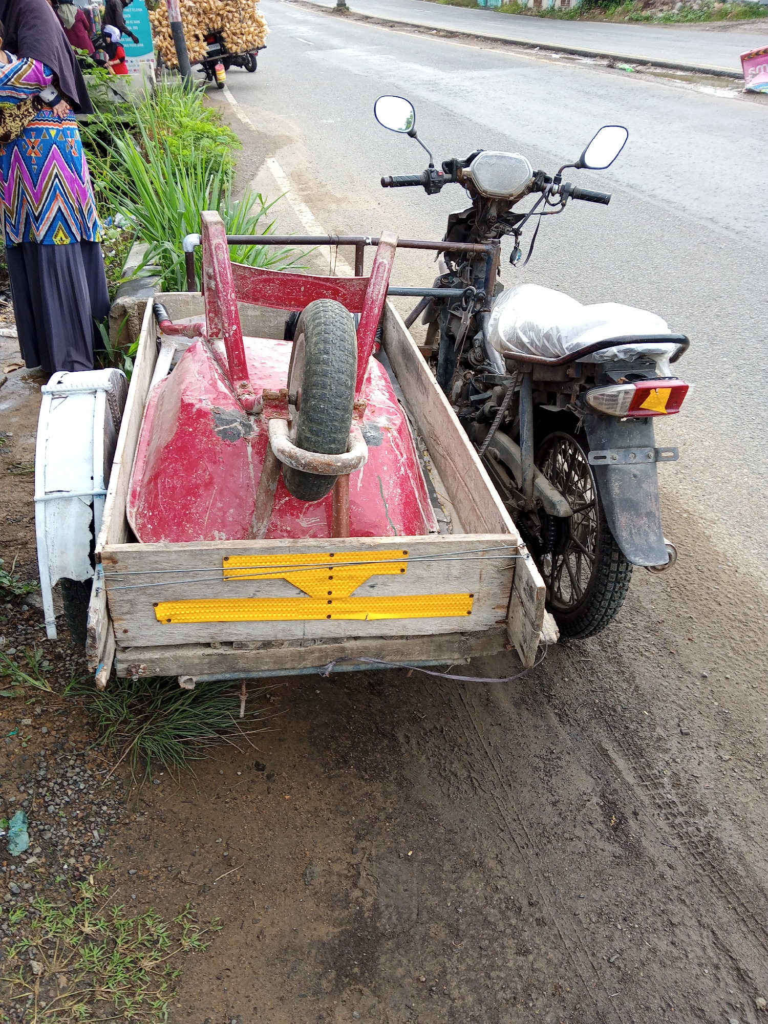 In Geulanggang Baro, Indonesia, a modified motorcycle with a sidecar is parked on the side of a paved road. The sidecar is constructed from wood and contains a red wheelbarrow. A person, partially visible on the left side of the image, is wearing a dark headscarf and a colorful patterned tunic over a dark skirt. Behind the motorcycle, a large pile of yellow produce, possibly corn, is loaded onto another motorcycle. The environment appears to be a rural or semi-urban area, with a dirt shoulder and some vegetation along the roadside. The weather is overcast.