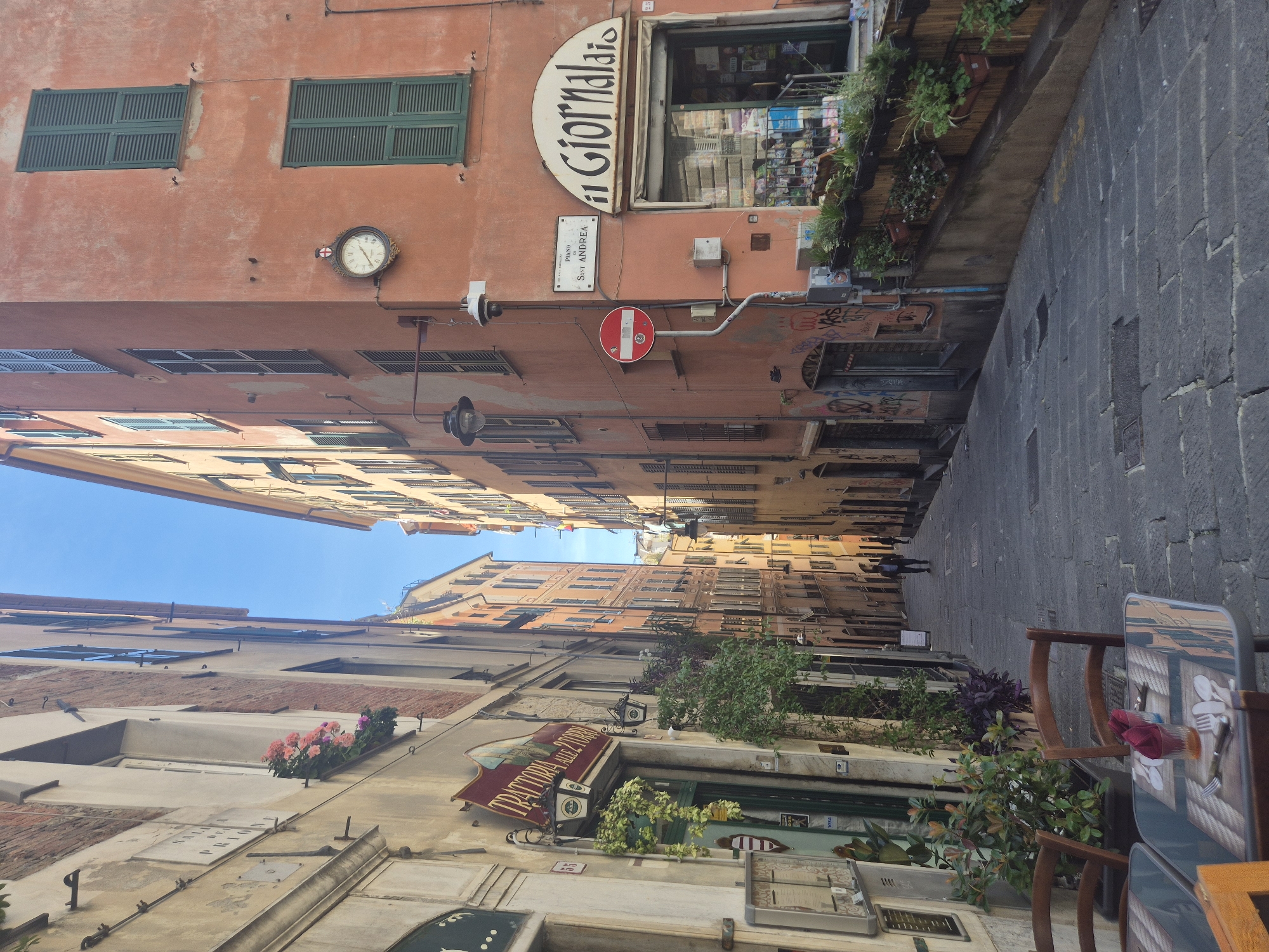 This is an eye-level, outdoor, daytime shot looking straight down a narrow street in Genoa, Italy. The perspective elongates the street, emphasizing the height of the buildings and the limited sky visible. Two people are walking far down the street. The tall, narrow buildings on either side feature various architectural details: windows with green shutters, flower boxes, and signage, including signs for "Il Giornalaio" and "Trattoria Alezzi 2008". A clock and a "Do Not Enter" sign are affixed to the upper portion of a building on the right. On the right side of the frame, a table and chairs suggest outdoor seating, with some utensils and napkins already on the table. The street is paved with dark gray cobblestones.