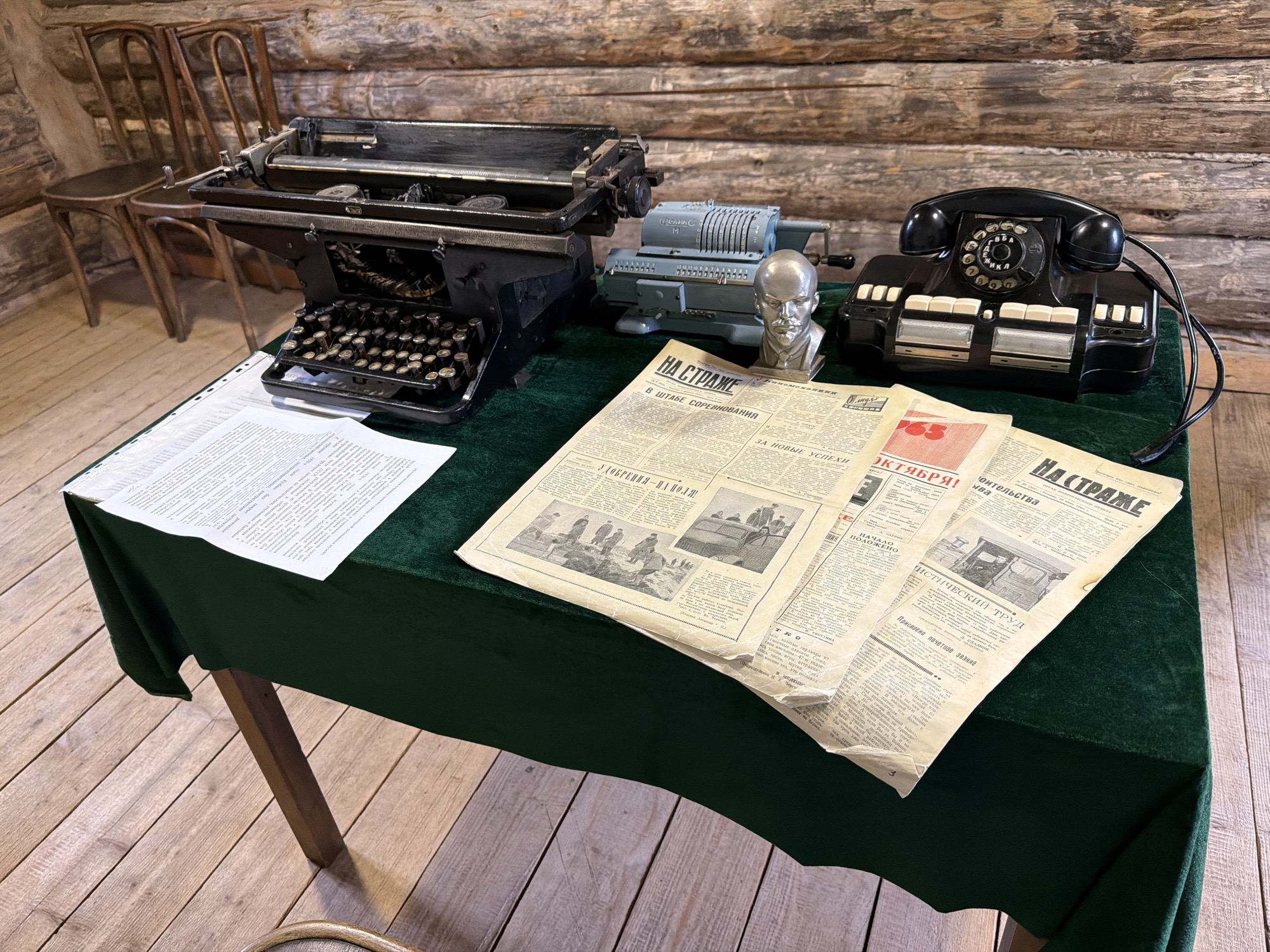 In a room with log walls and a wooden floor, a dark green tablecloth covers a table. On the table are several objects: a black manual typewriter, a light blue mechanical calculator, a silver bust of Lenin, and a black rotary telephone. Scattered across the table are newspapers. Two wooden chairs are visible in the background. The scene appears to be a static display, possibly in a museum or historical exhibit, depicting a historical office setting. The city of Ust'-Kabyrza, Russia is a rural settlement, and this scene may represent a historical office from that region or a similar setting.