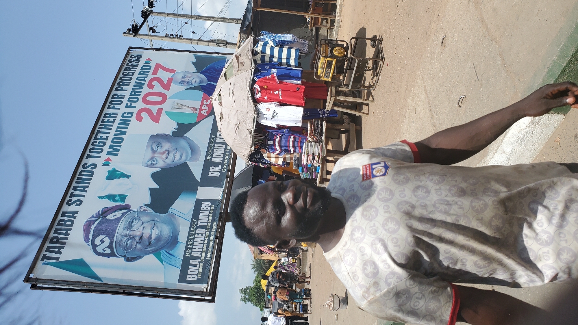 This image captures a street scene in Jalingo, Nigeria, with a large billboard dominating the upper portion. The billboard features portraits of two men, Bola Ahmed Tinubu, identified as President, Commander-in-Chief of The Armed Forces, Federal Republic of Nigeria, and Dr. Agbu Kefas, Governor Taraba. The text on the billboard reads "TARABA STANDS TOGETHER FOR PROGRESS MOVING FORWARD 2027."
In the foreground, a man with dark skin and a beard is looking to the side, smiling slightly. He is wearing a light-colored t-shirt with a repeating pattern and a logo on the left chest. Behind the man, a market stall is visible, with clothing items hanging and stacked on tables. These include various t-shirts, some with sports branding. A yellow generator is placed on the ground near the stall. In the background, other people and structures suggest a busy urban environment. The sky is clear and blue, indicating daylight.