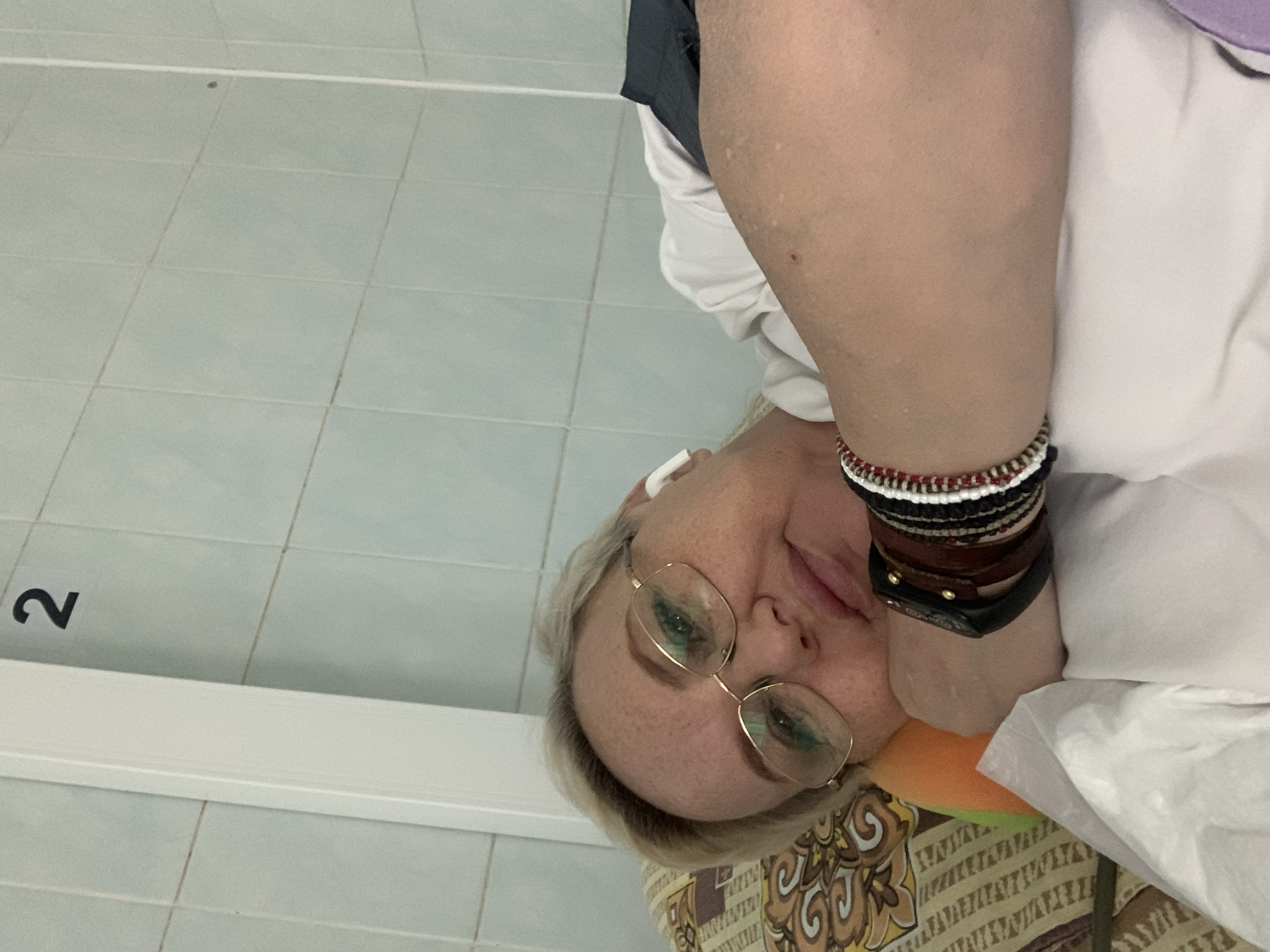 A woman is depicted in a close-up, lying or reclining indoors, with her head resting on a patterned cushion featuring brown, yellow, and orange designs. She has short, light blonde hair and wears gold-rimmed glasses, through which her green eyes are visible. A white earbud is positioned in her right ear, and she appears to be looking towards the camera with a calm and slightly smiling expression. Her left arm, adorned with a collection of bracelets and bands including colorful beaded ones, woven cords, leather straps, and a dark-colored fitness tracker or watch, is visible. She is dressed in a white shirt, and a small, vibrant orange and green object is tucked beneath her chin, likely serving as a neck support or small cushion.
The setting appears to be an indoor space with light blue-green square tiles forming the background, indicative of a clean, possibly institutional environment such as a hospital, clinic, or waiting area in Chaikovsky, Russia. A prominent black number "2" is visible on one of the tiles to the left of the woman. Below this, a stark white, horizontal panel or structure runs across the frame. The lighting is uniform, suggesting artificial indoor light. Small, faint, lighter spots are visible on her arm, along with a tiny reddish blemish. The image captures a moment of rest or quiet contemplation in this internal space.