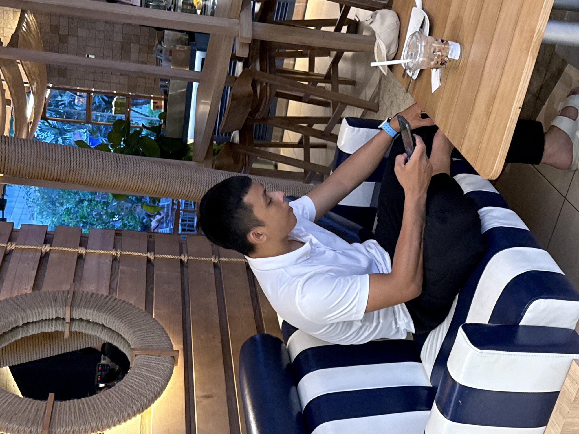 A young Vietnamese man in a white polo shirt and black pants sits on a blue and white striped couch in a cafe. He is looking at his phone and wearing a blue watch. On the wooden table in front of him is a plastic cup with a drink, a straw, and a white cap. In the background, there are wooden chairs and tables, and greenery visible through a window. The cafe has a rustic design with wooden walls and rope accents. This scene is likely located in Nha Trang Ward, Vietnam.