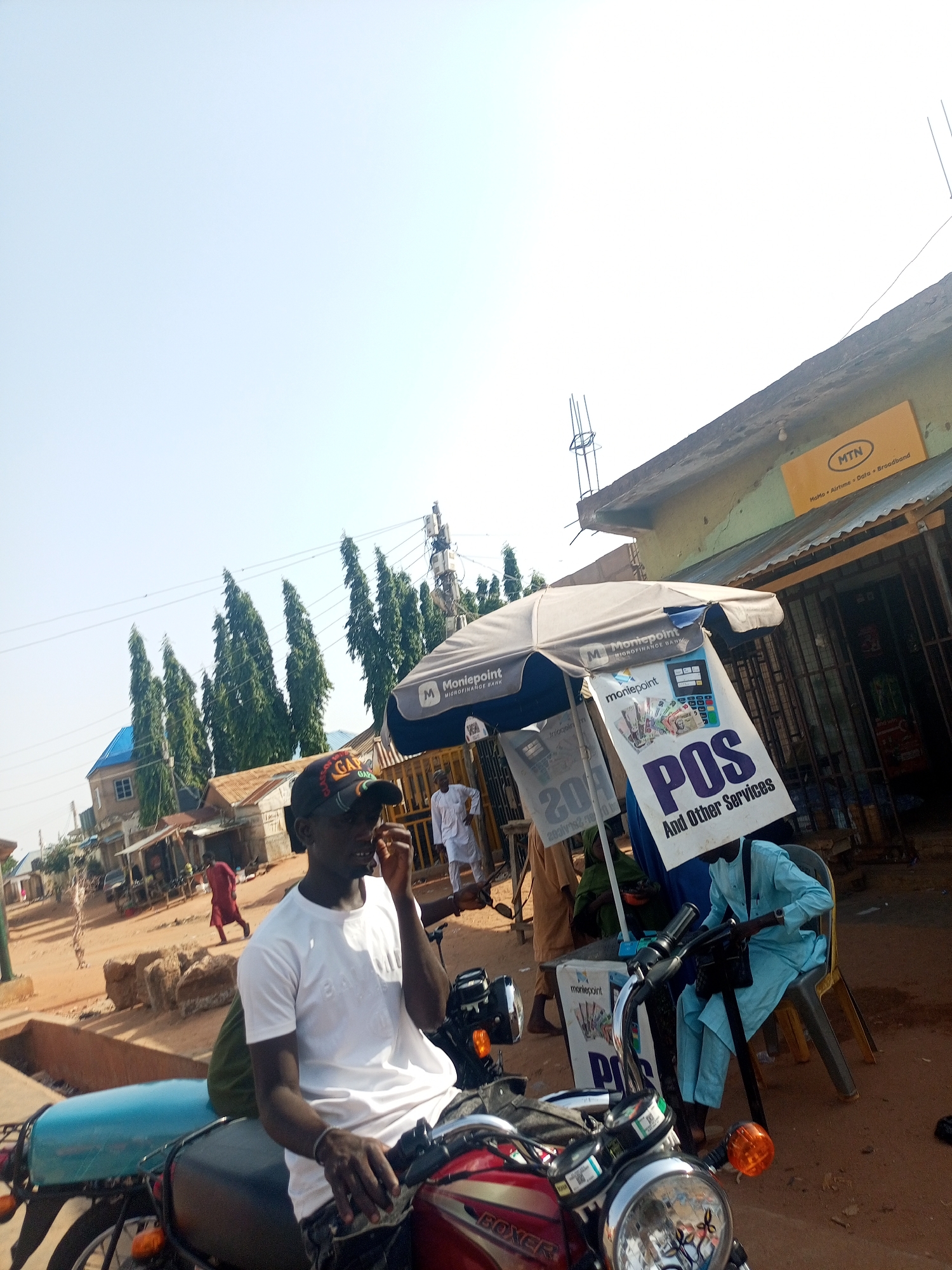 The image shows a street scene in Gombe, Nigeria. A man wearing a white t-shirt and a baseball cap is sitting on a red Boxer motorcycle. Next to him is a Point of Sale (POS) station under a Moneypoint umbrella, operated by a man wearing a light blue outfit. A sign advertises "POS And Other Services." In the background, there are buildings, trees, and other people walking along the street. An MTN sign is visible on one of the buildings. The environment appears to be a typical bustling street in a Nigerian city.