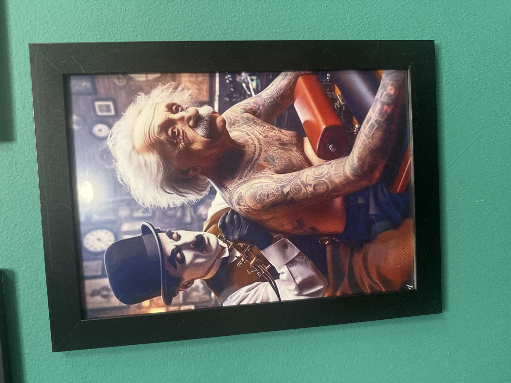 The image shows a framed picture on a teal-colored wall. The picture depicts Albert Einstein with numerous tattoos on his chest and arms, sitting on a tattoo chair. Charlie Chaplin, wearing his signature bowler hat and makeup, is tattooing Einstein. The background of the picture appears to be a dimly lit room with clocks on the wall. There is no information in the image to suggest any connection to Yegorlykskaya, Russia.