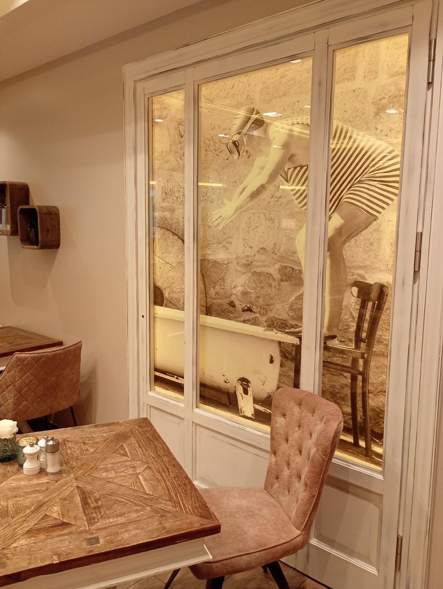 This image captures a scene inside a cafe or restaurant, specifically located in Bad Sachsa, Germany. The foreground features a wooden table with a decorative inlay pattern, set with salt and pepper shakers and a small vase with a white flower. A tufted, rose-colored chair is positioned next to the table.
The background is dominated by a large glass-paned door or window. Through the glass, a vintage-style black and white photograph is displayed, depicting a person wearing a striped bathing suit and swim cap in a diving pose. Behind the photographic display, a white bathtub and a wooden chair are visible, along with a stone wall. Two small, square wooden shelves are mounted on the light-colored wall to the left of the display. The overall ambiance suggests a retro or themed interior design.