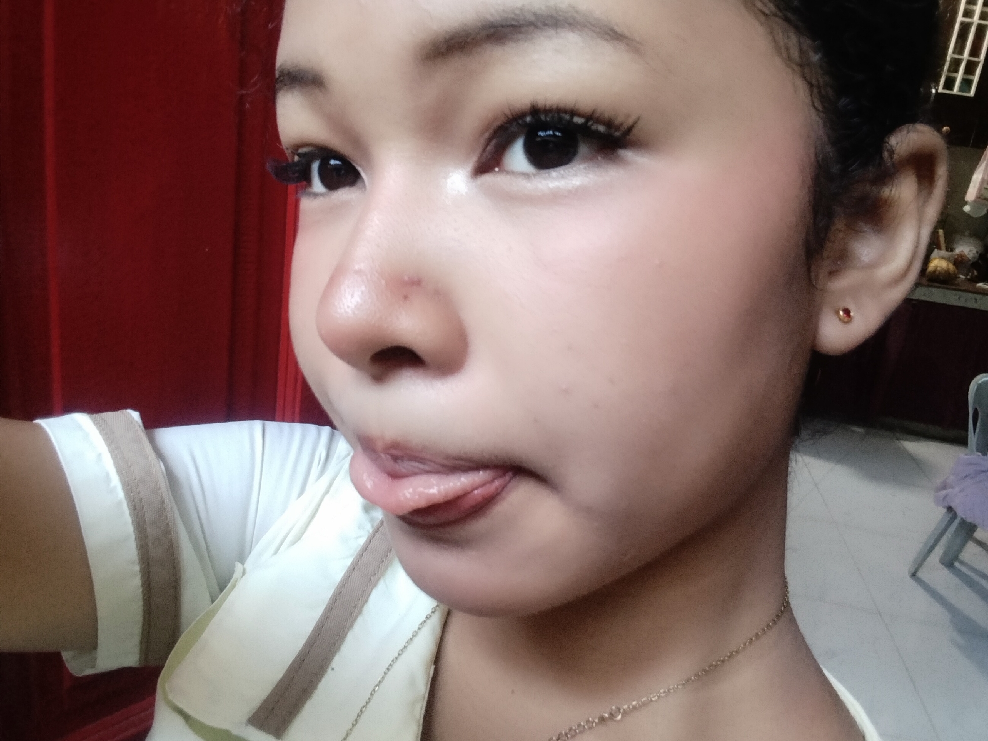 A close-up, low-angle selfie captures the face of a young woman with dark hair, styled in a bun, and fair skin. She is wearing a white top with tan trim and a delicate gold necklace. Her eyes are open, and she is sticking her tongue out. Her makeup includes mascara and blush. A small gold earring with a red gem is visible in her right ear. The background shows a red curtain on the left and a blurry interior scene on the right, including a chair with a light purple cloth. The lighting is diffused, creating a soft glow on her face. The image was likely taken indoors in a residential setting.