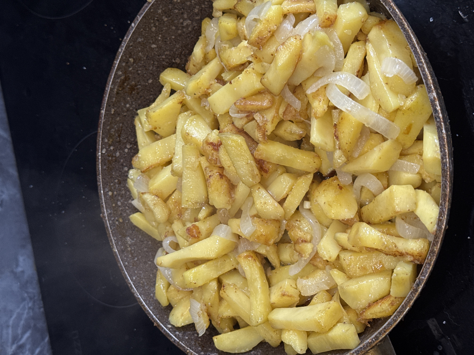 The image shows an overhead view of fried potatoes and onions in a dark frying pan. The pan, which appears to have a speckled, possibly non-stick interior and a slightly worn edge, is positioned on a dark, glossy stovetop.
The primary subjects are the food items: numerous pieces of golden-brown fried potatoes cut into irregular, elongated shapes, mixed with translucent white slices of onion. Some potato pieces exhibit darker, slightly crispy-looking edges, while the onions are soft and integrated throughout the dish. The food has a noticeable sheen, indicating it has been cooked with oil or fat.
The activity depicted is the cooking or the resting of cooked food in a pan on a stovetop. The dark stovetop surface shows faint circular outlines, characteristic of an electric or induction hob. A small portion of a lighter, possibly marble or tiled surface, is barely visible in the top-left corner, suggesting a kitchen setting. There are no people visible. No discernible text is clearly visible on the pan or the stovetop. The lighting is bright and even, highlighting the texture and color of the food.
