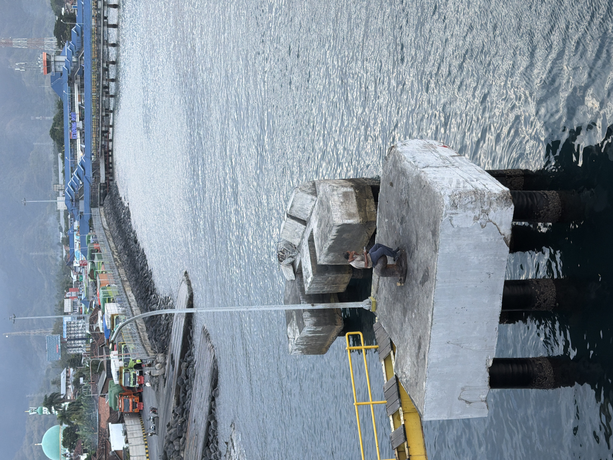 The image depicts a view of Ketapang Port in Ketapang, Indonesia. In the foreground, a man is seated on a bollard on a concrete pier, appearing to be looking at a phone. The pier is supported by dark, barnacle-covered pillars extending into the water. Adjacent to the pier is a yellow railing and a wooden walkway.
In the middle ground, a breakwater made of large rocks lines the edge of the port. Beyond the breakwater, a paved area is visible with several trucks lined up, suggesting port activity. Buildings, including a green-domed structure resembling a mosque and other industrial or commercial buildings, are situated along the coastline. A tall lamppost extends upwards from the pier.
The background shows a body of water, likely the sea, with gentle ripples. In the distance, a larger port facility with blue-roofed buildings and a control tower is visible. Hills or mountains form the backdrop under an overcast sky. The overall scene conveys a working port environment with elements of local architecture and infrastructure.