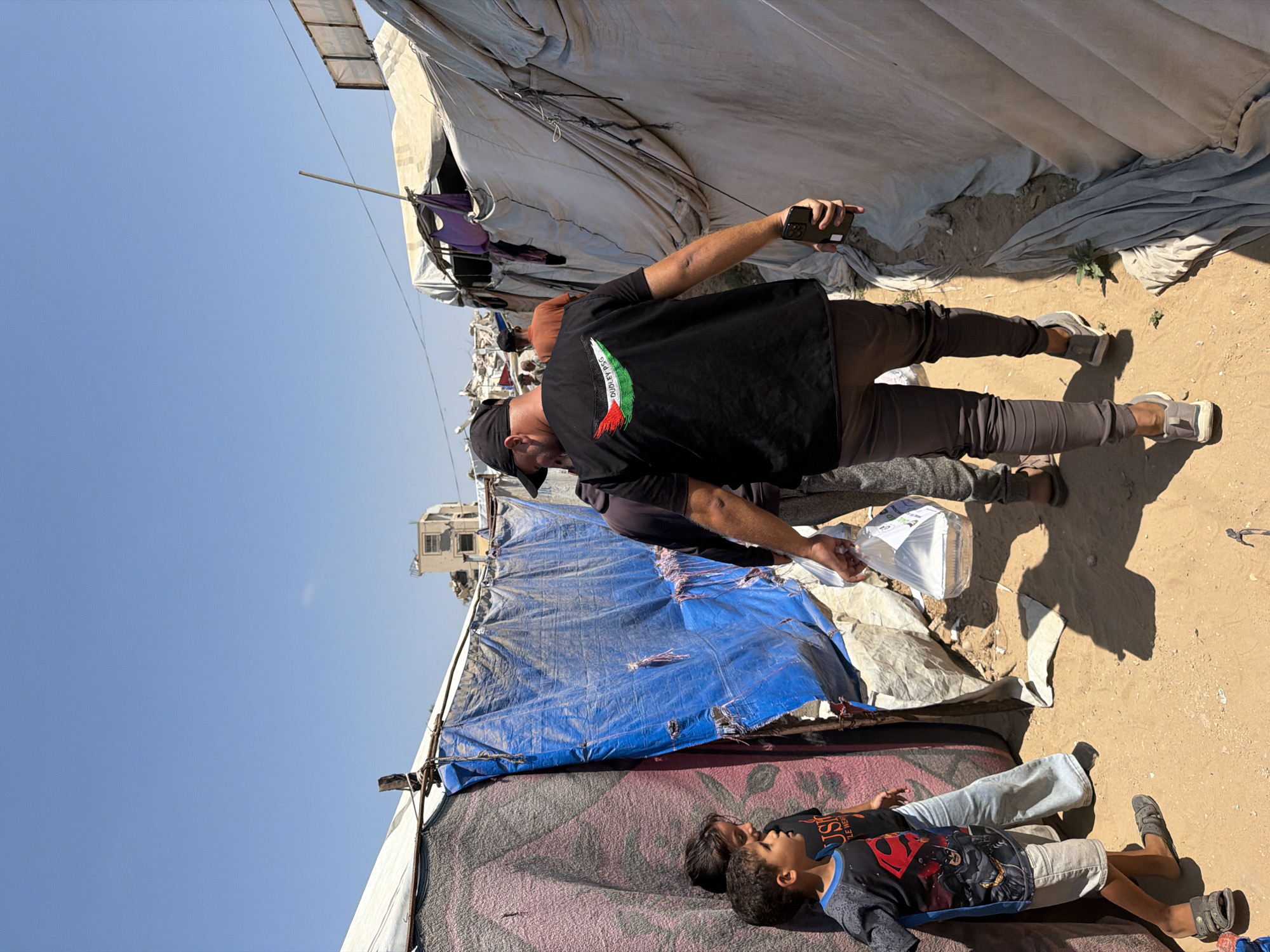 In Jabalia, Palestinian Territories, under a clear blue sky, a man wearing a black vest with a Palestinian flag emblem and the words "DUDLEY PSG" walks away from the camera down a narrow dirt path. He is carrying a foil food container in a clear plastic bag in his left hand and holding a smartphone in his right. To his left, two children stand near a makeshift shelter constructed from tarps and other materials. One child, wearing a dark t-shirt with a superhero graphic, looks towards the path. The other child, partially obscured, also faces the path. The surrounding environment appears to be a densely populated area with temporary structures, suggesting a refugee camp or informal settlement. In the background, damaged buildings are visible, indicating past destruction. The scene depicts an act of aid distribution in a community affected by conflict.