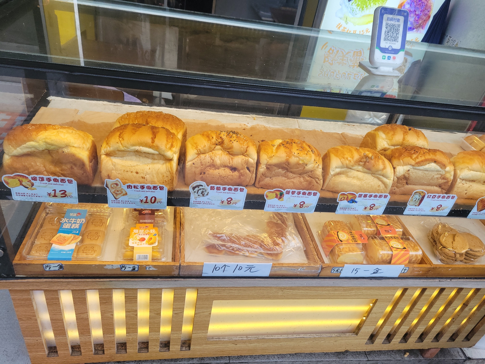 The image captures a close-up view of a glass display case filled with a variety of baked goods, likely in a bakery or food stall in Liangjiang, China. The scene focuses on the products offered for sale.
The top shelf features several loaves of hand-shredded bread (手撕面包) with different fillings. From left to right, visible types include Durian Hand-Shredded Bread (榴莲手撕面包) for ¥13, Pork Floss Hand-Shredded Bread (肉松手撕面包) for ¥10, and several varieties like Grape (葡萄), Pineapple (菠萝), Blueberry (蓝莓), and Red Bean (红豆) hand-shredded bread, all priced at ¥8. Most loaves are golden brown, some topped with sesame seeds.
The bottom shelf displays pre-packaged snacks. On the left are boxes of "Buffalo Milk Cake" (水牛奶蛋糕) from the "Mengbao" (梦堡) brand, priced at 12 yuan. Adjacent to these are "Bursting Small Soft Cakes" (爆浆小软) in molten chocolate and molten egg yolk flavors, with a visible sign stating "10个10元" (10 pieces for 10 yuan). Further right are "Tiger Skin Roll Cakes" (虎皮卷蛋糕) priced at ¥8, and a bag of round, flat cookies.
In the background, visible through the glass, is a white QR code payment stand labeled "扫码付款" (Scan code to pay), displaying "抹茶红豆 15元" (Matcha Red Bean 15 yuan) and a customer service number (400-919-6688). The lower part of the display case is made of wooden slats with internal illumination, suggesting an evening or indoor setting, though reflections on the glass hint at an exterior environment. No people are visible, emphasizing the products for sale.
