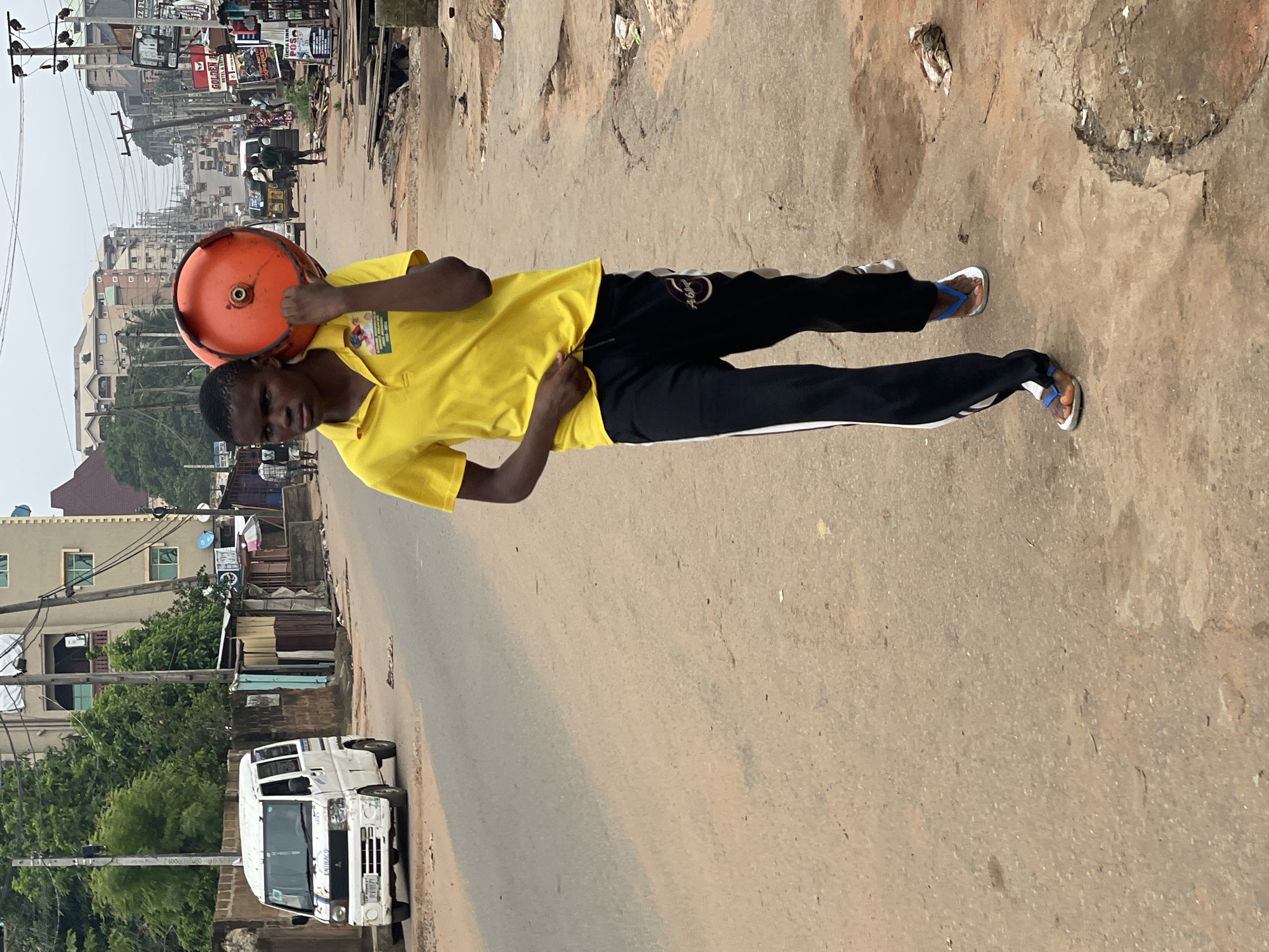 A medium shot captures a young man walking along a road in Nkpor, Nigeria. He is carrying an orange gas cylinder on his left shoulder. The man wears a yellow polo shirt and black pants with a white stripe. He is wearing blue and white flip-flops. The environment around him is a typical street in Nkpor, with buildings, shops, and utility poles visible in the background. A white van is parked on the side of the road. The road itself is paved but appears worn.