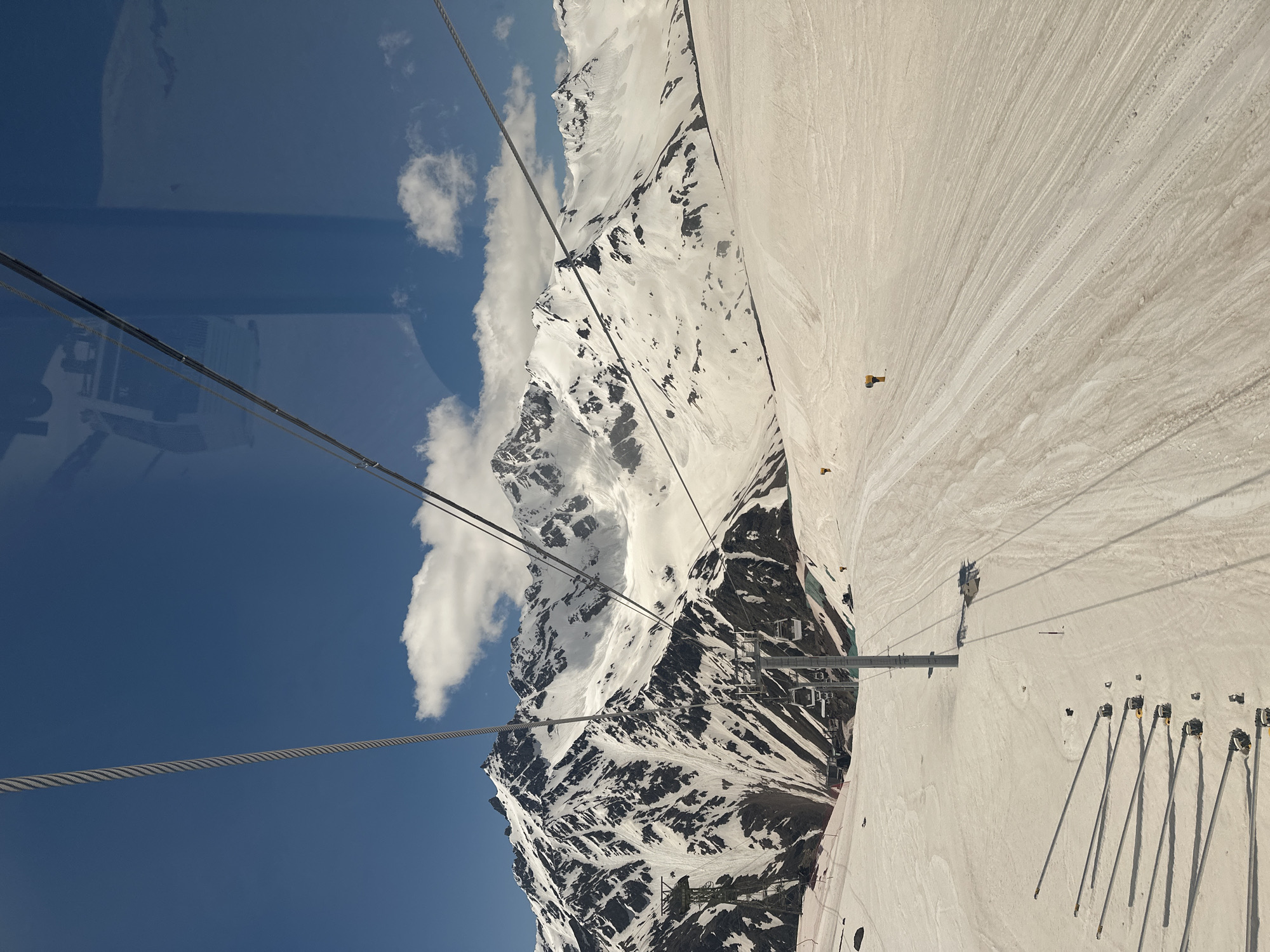 This image depicts a ski resort scene in Terskol, Russia, captured from the perspective of a ski lift. In the foreground, ski tracks are visible on a snow-covered slope, with several poles casting long shadows. A ski lift with multiple cabins is ascending the mountain. The background features majestic, snow-capped mountains under a clear blue sky dotted with white clouds. The scene is brightly lit by sunlight.