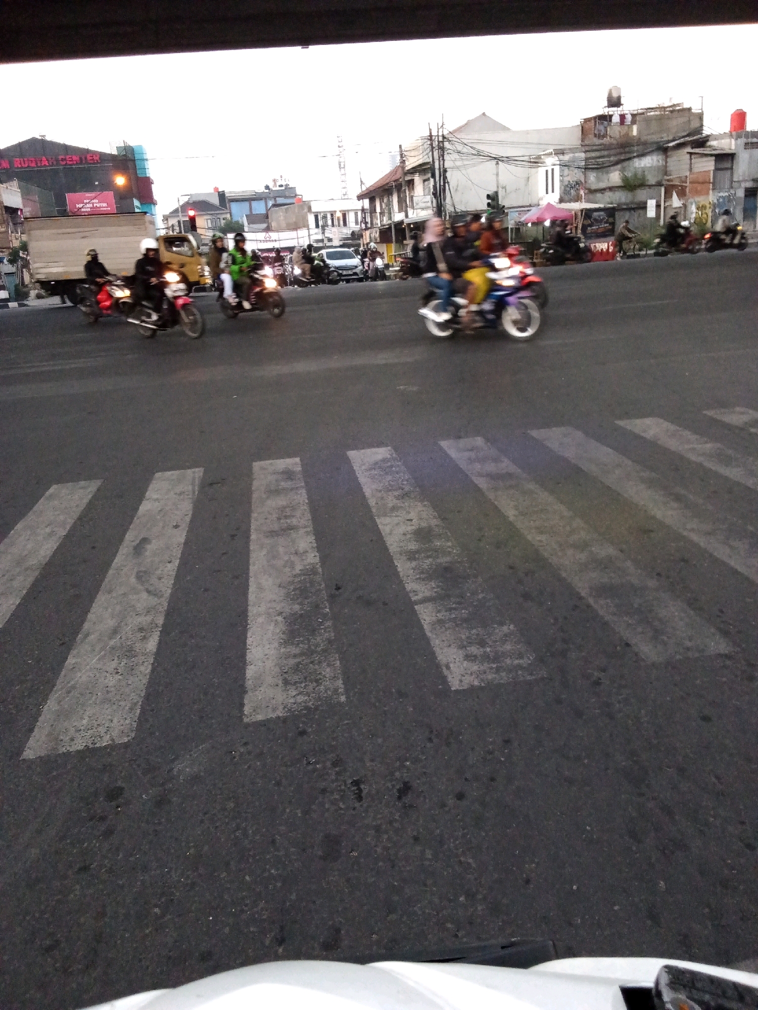 The image captures a street scene in Bandung City, Indonesia, likely at an intersection with a pedestrian crossing. Multiple motorcycles with riders are visible on the asphalt road, some of them in motion. In the background, buildings line the street, including a structure with prominent signage that appears to read "RM RUJIAH CENTER." There is a delivery truck parked on the left side of the road. The sky is overcast, suggesting it might be late afternoon or early morning. The foreground is dominated by a pedestrian crossing with white stripes painted on the dark asphalt. The overall impression is that of an active urban environment with vehicular traffic.